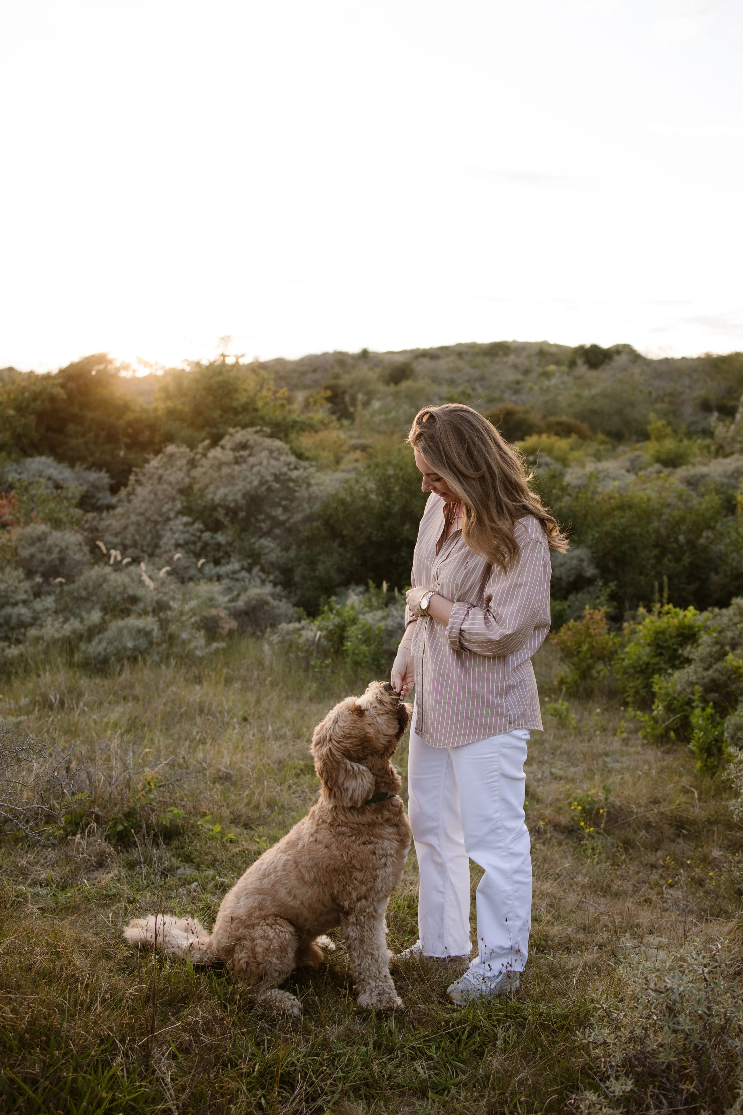 Een vrouw in een gestreepte blouse en witte broek staat in een groene omgeving, terwijl ze liefdevol naar haar beige hond kijkt die aan haar voeten staat, with the ondergang van de zon op de achtergrond.