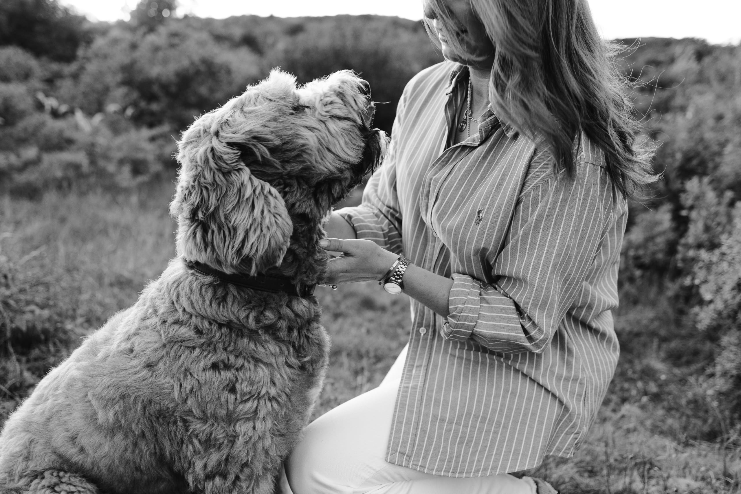 Een vrouw knielt in een groene omgeving terwijl ze liefdevol naar haar grote, harige hond kijkt, die haar aandacht en genegenheid toont. De foto is in zwart-wit, wat de emotionele band tussen hen benadrukt.