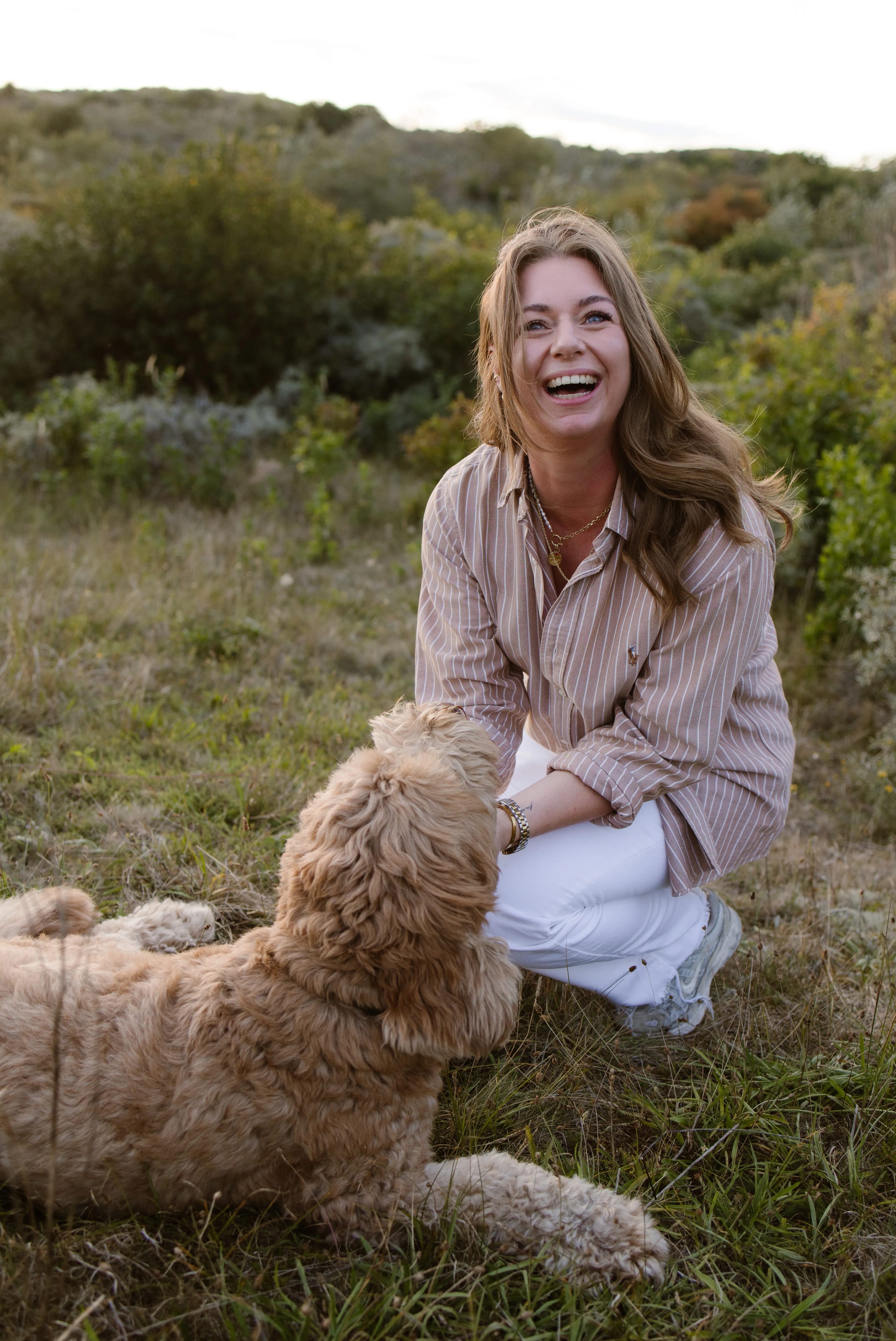 Een vrouw in een gestreepte blouse lacht terwijl ze met een grote, schattige hond in een groene, natuurlijke omgeving op een grasveld speelt.