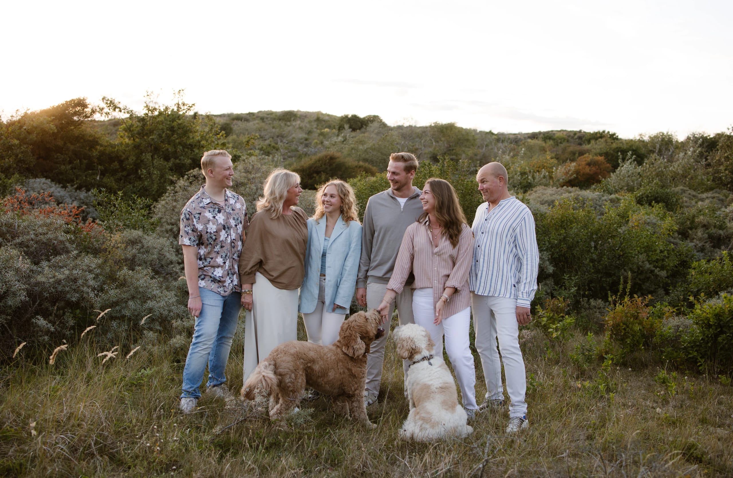 Een gelukkige familie staat samen in een groene weide, omringd door bomen en struiken, met twee honden aan hun voeten, terwijl de zon ondergaat in de achtergrond.