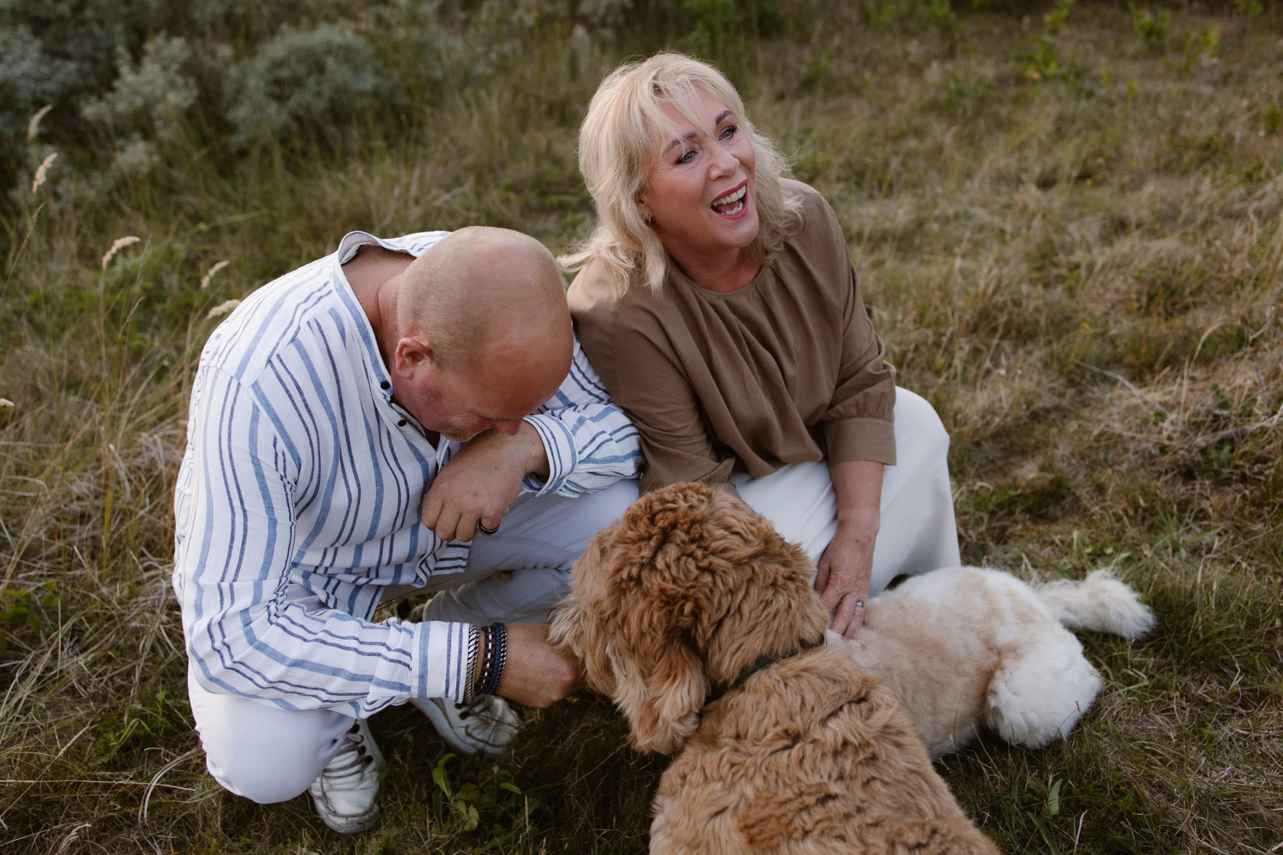 Een vrolijk stel zit op een grasveld, waarbij de vrouw lacht terwijl ze naar hun hond kijkt, terwijl de man zich gebogen heeft en liefdevol met de hond speelt. De rustige natuurscène straalt een gevoel van verbondenheid en vreugde uit.