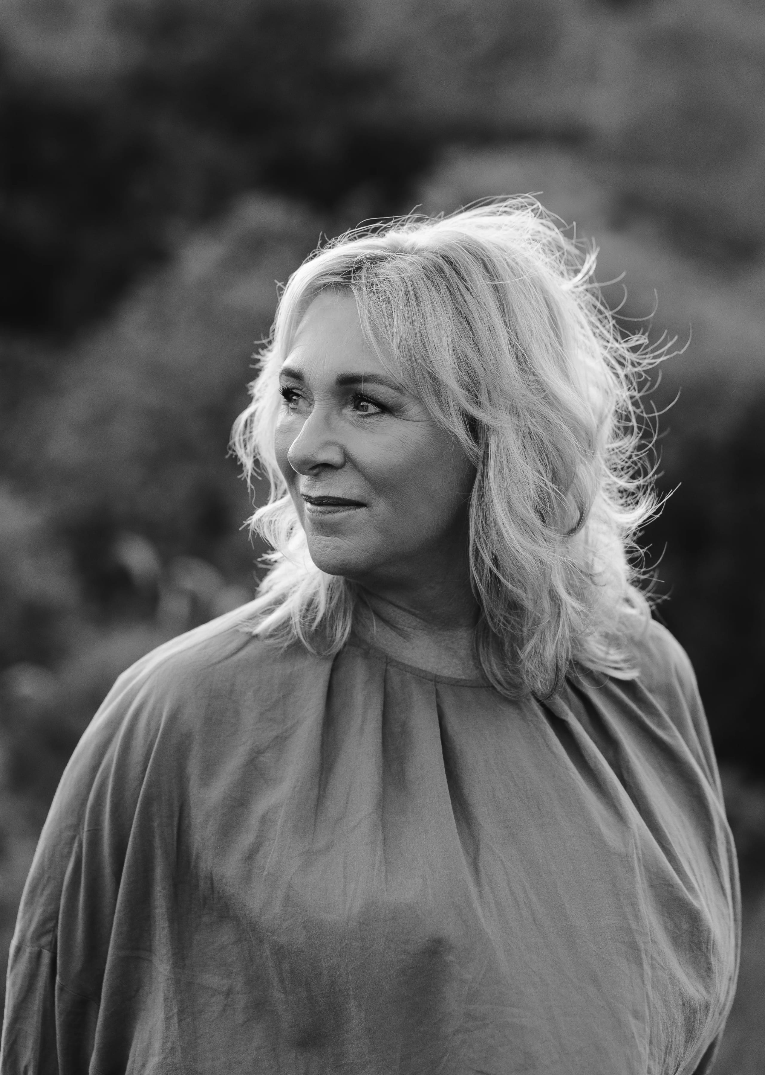 Een vrouw met krullend, blond haar staat in een natuurlijke omgeving en kijkt contemplatief opzij, gekleed in een elegante, losse blouse. Het zwart-wit beeld straalt een gevoel van rust en zelfreflectie uit.
