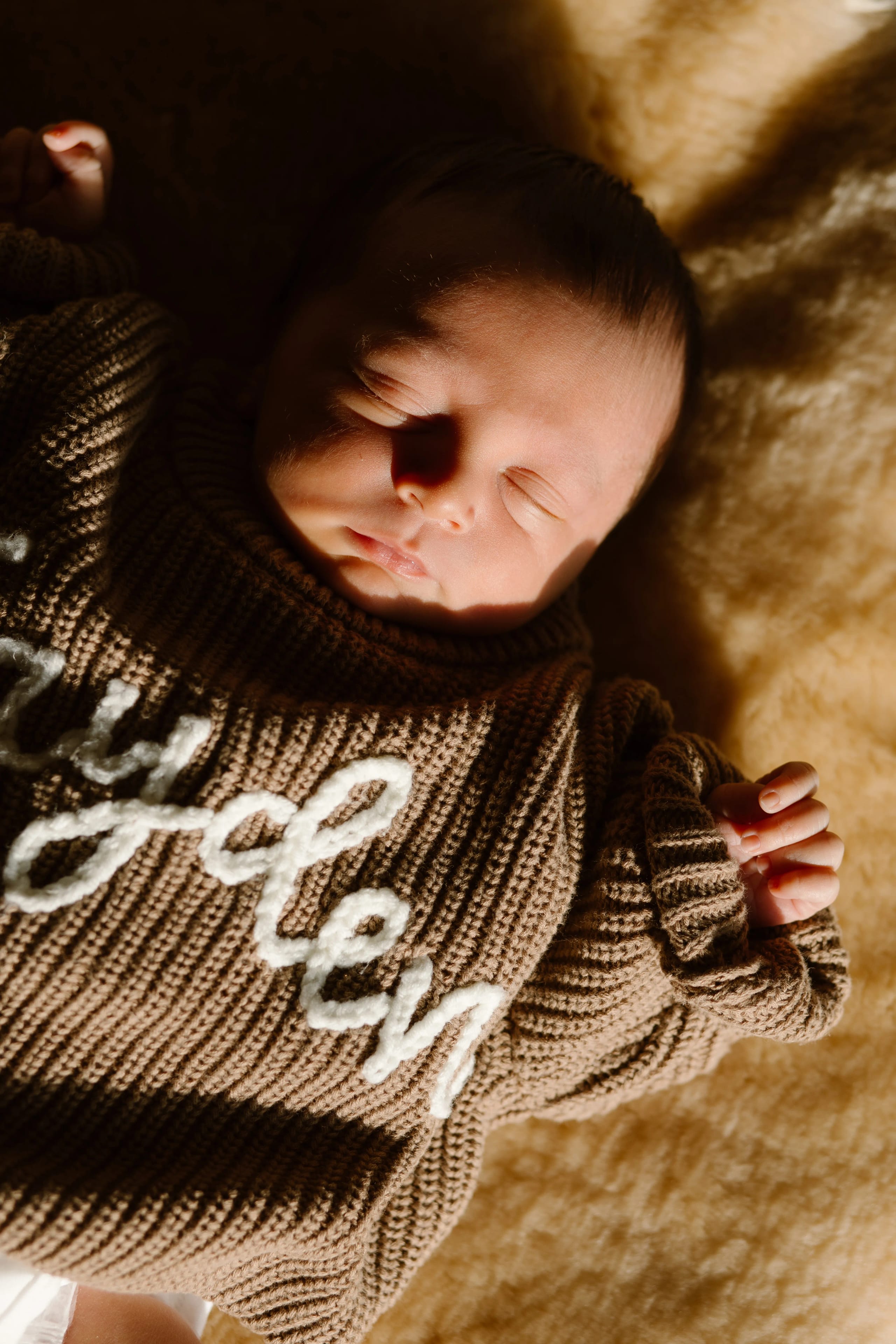 Slapende baby in een zachte, beige trui met gebreide textuur en de naam 'Jayden' in witte letters, rustend op een warme ondergrond met natuurlijk licht.