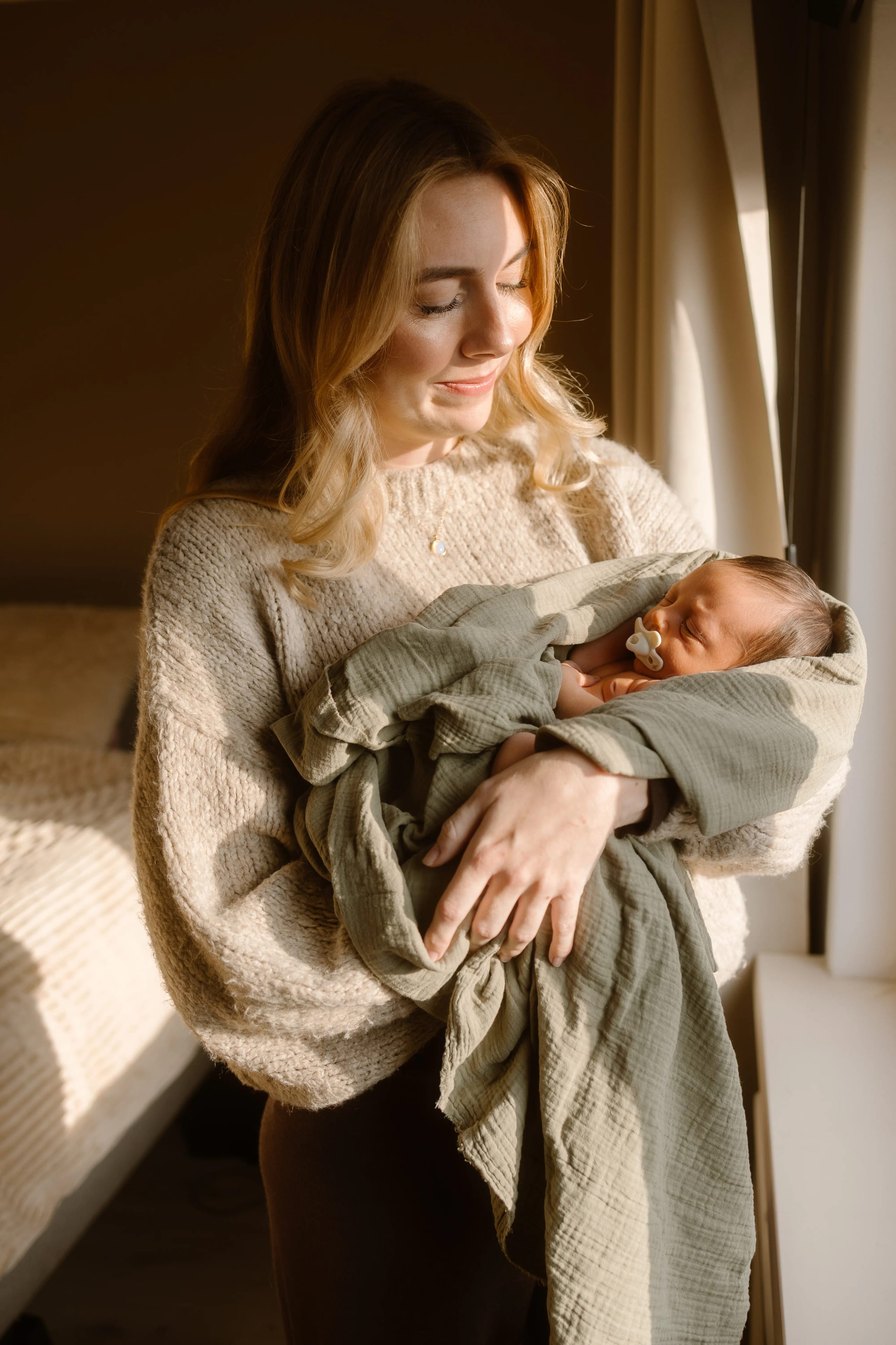 Een jonge vrouw houdt een pasgeboren baby in haar armen, omhuld in een groene deken, terwijl haar gezicht een liefdevolle glimlach toont, met natuurlijk licht dat door het raam valt en een warme sfeer creëert.
