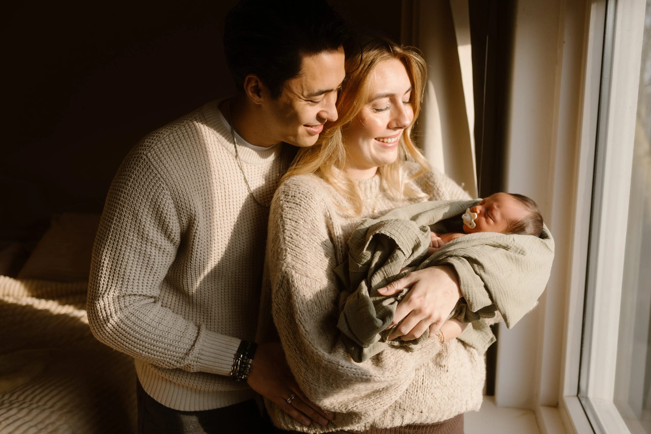 Een blijdschap uitstralend stel staat dicht bij elkaar in een sfeervol verlicht interieur, terwijl ze samen hun pasgeboren baby vasthouden, liefdevol ingepakt in een groene dekentje.