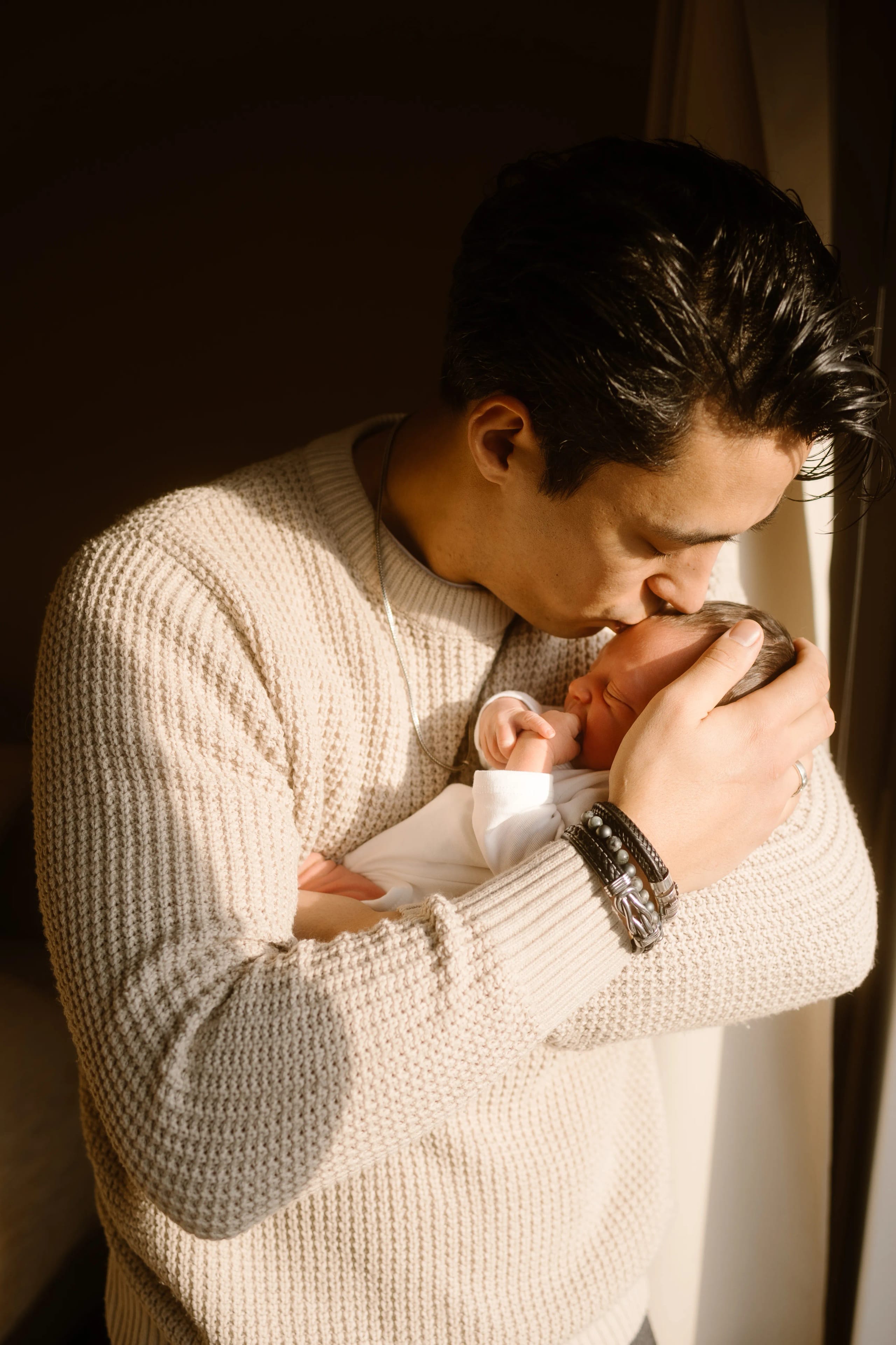 Een vader kust voorzichtig zijn pasgeboren baby terwijl hij hem in zijn armen houdt, omringd door warm natuurlijk licht dat door het raam valt.