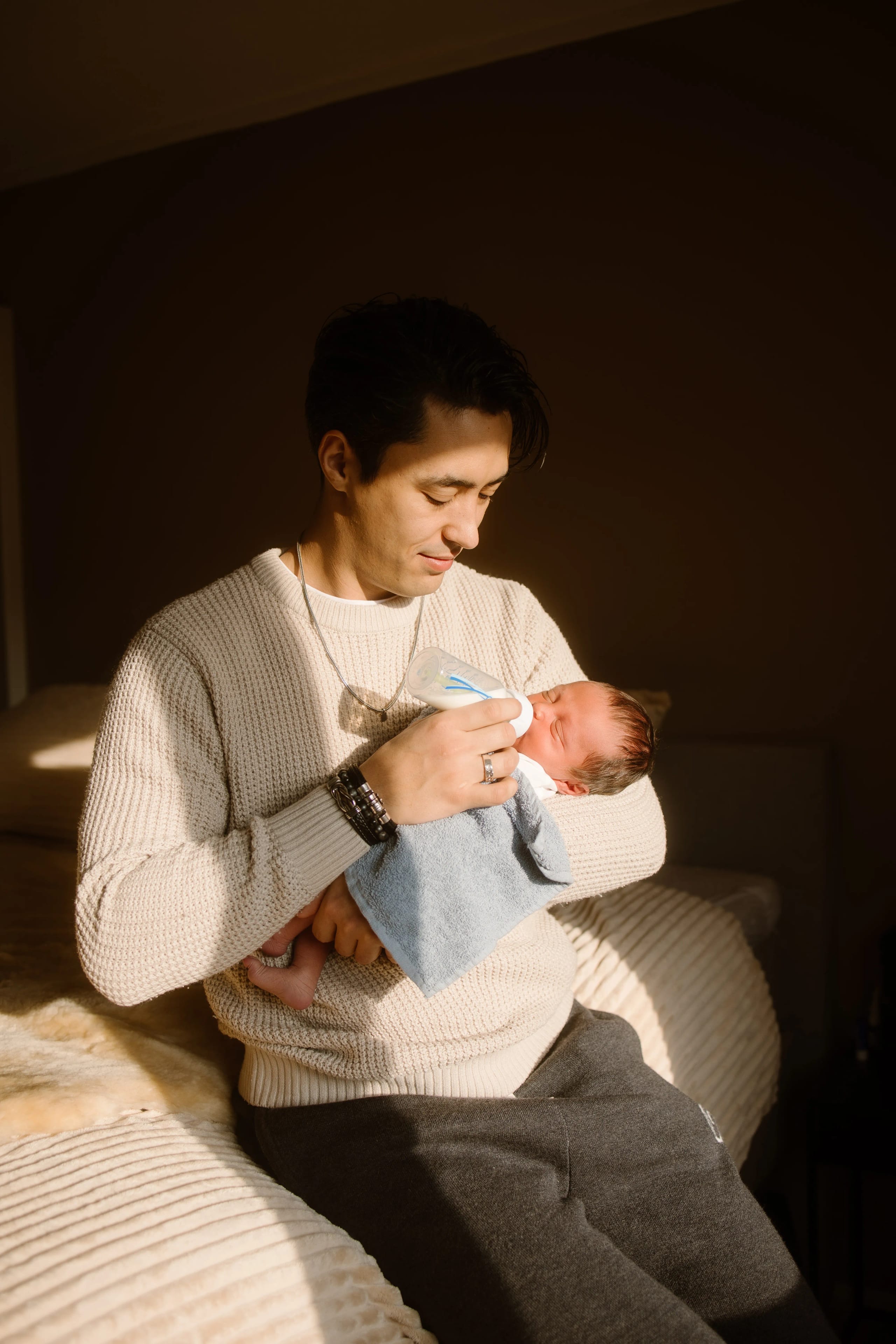 Een jonge vader voedt zijn pasgeboren baby met een fles terwijl hij op een bed zit, omringd door zacht licht dat binnenkomt vanuit een raam.