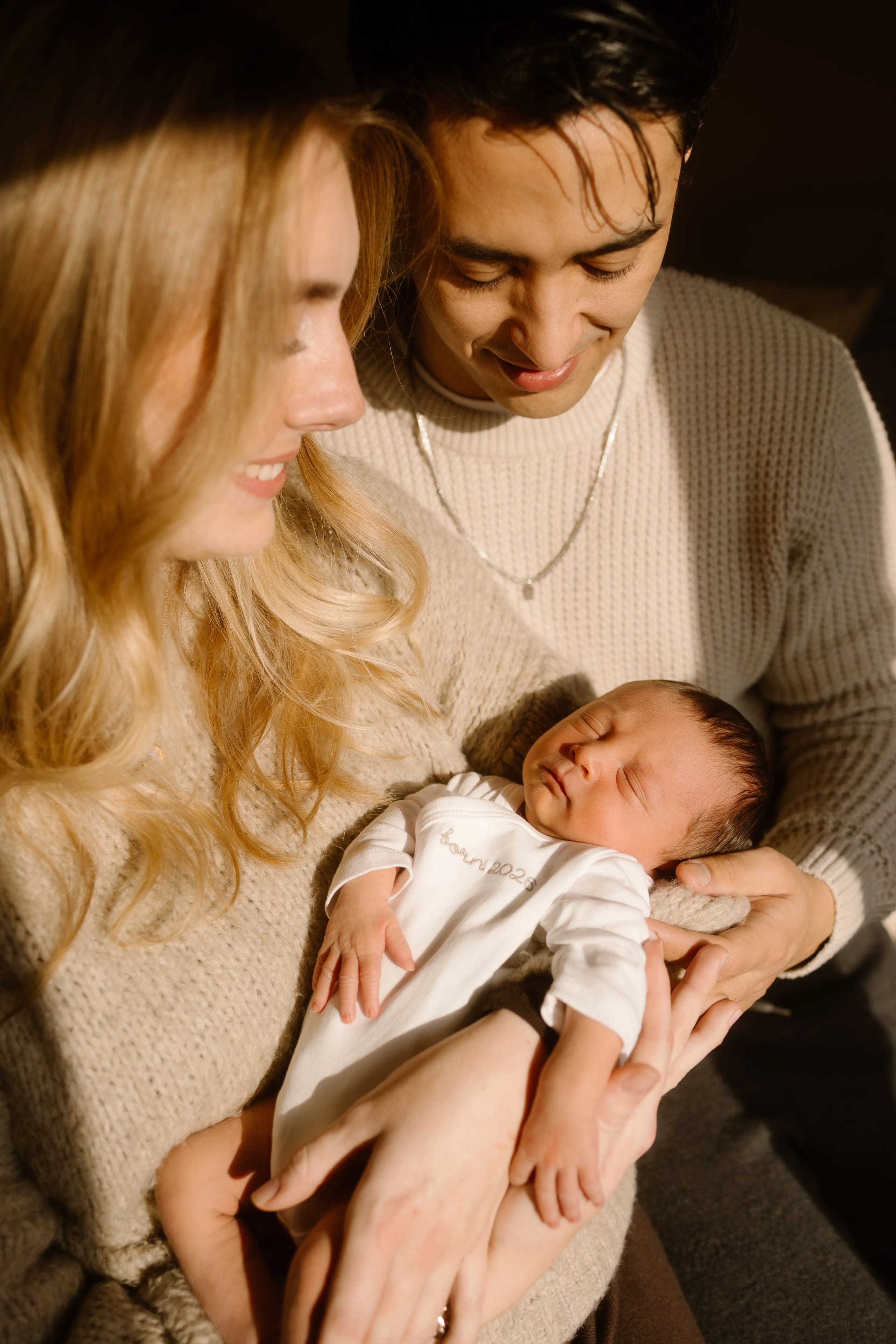 Gelukkige ouders houden hun pasgeboren baby vast, terwijl warme zonnestralen hun liefdevolle momenten verlichten, met de baby gekleed in een schattige witte romper.