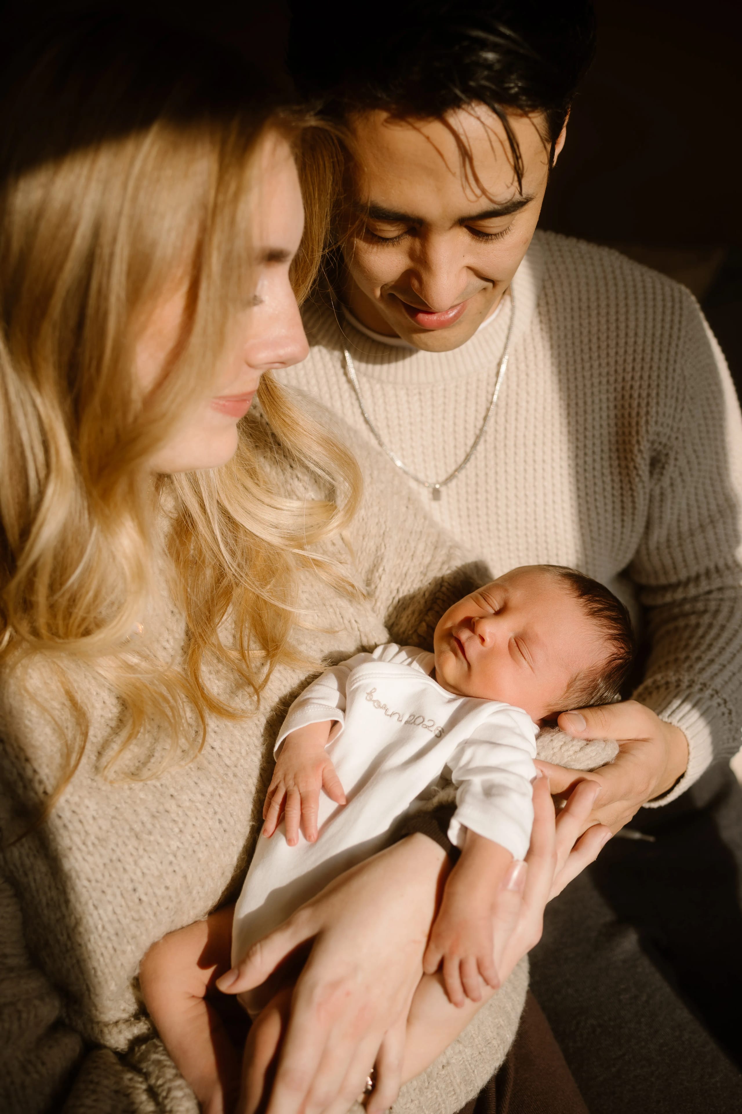 Een blije nieuwe ouders houden hun pasgeboren baby vast in hun armen, terwijl ze genietend naar het kindje kijken, omgeven door zacht, natuurlijk licht dat de intieme sfeer van het moment benadrukt.