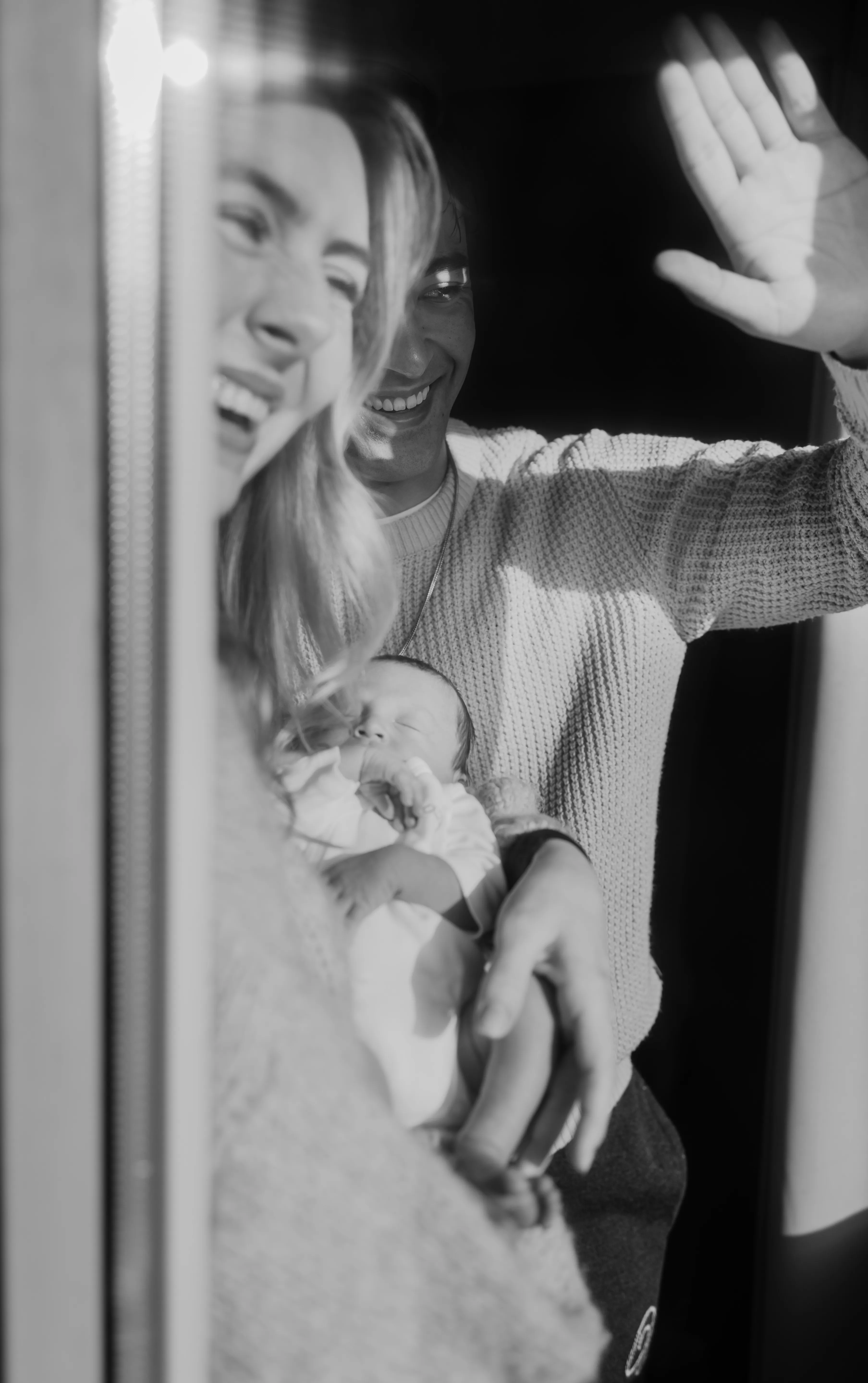 Een gelukkige jongeman en vrouw lachen terwijl ze vanuit een raam met de hand groeten, terwijl de vrouw een pasgeboren baby in haar armen houdt. De sfeer is warm en liefdevol, met een heldere lichtinval die de vreugde van het moment benadrukt.