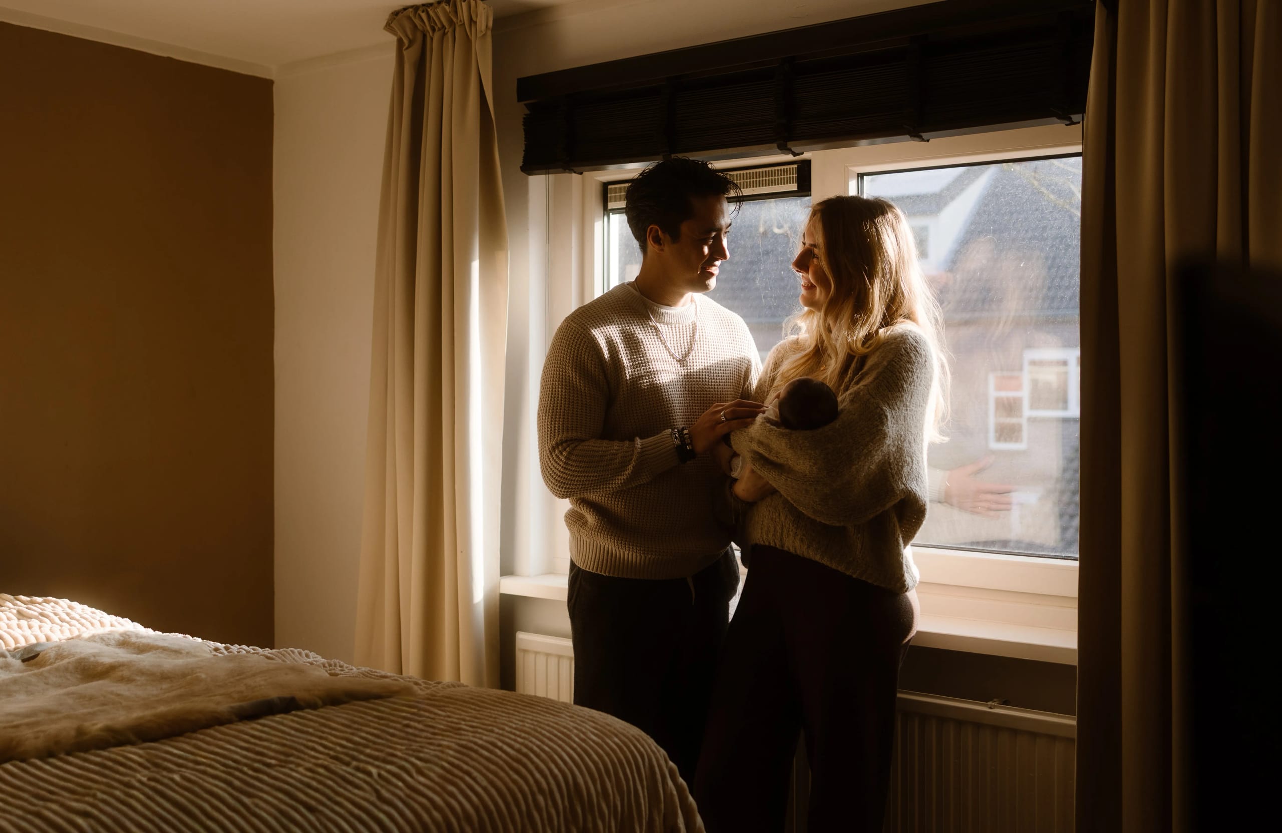 Een gelukkig stel staat bij een raam in een sfeervol verlichte slaapkamer, terwijl ze samen hun pasgeboren baby bewonderen. Zachtjes omarmd en met liefdevolle blikken, creëren ze een intieme en warme sfeer in hun huiselijke omgeving.