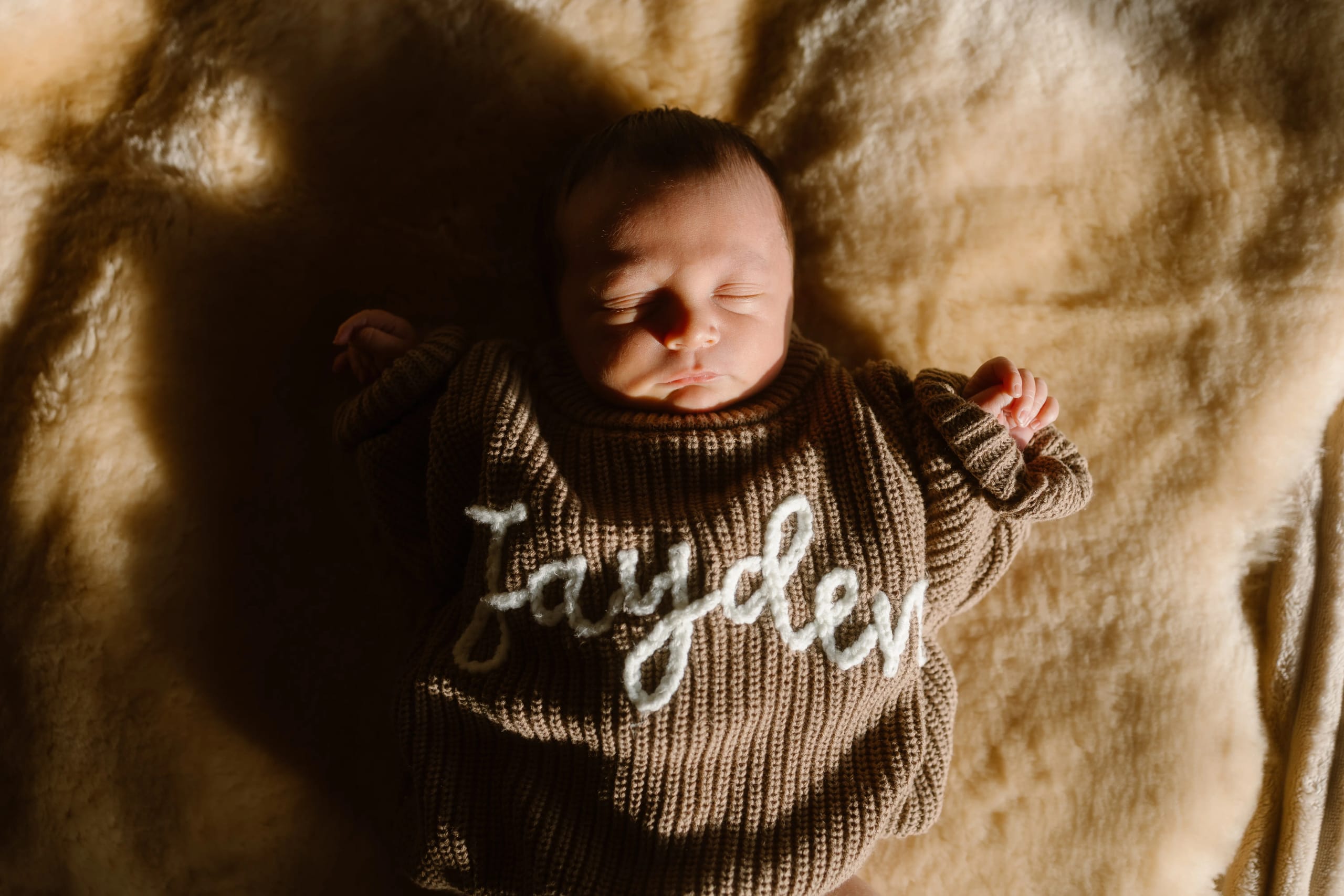 Pasgeboren baby ligt rustig en vredig op een zachte, lichte ondergrond, gekleed in een bruin gebreid truitje met de naam "Jayden" in witte steken. Het warme licht benadrukt de schattige gelaatsuitdrukking en de kleine, opgerolde handjes van de baby.