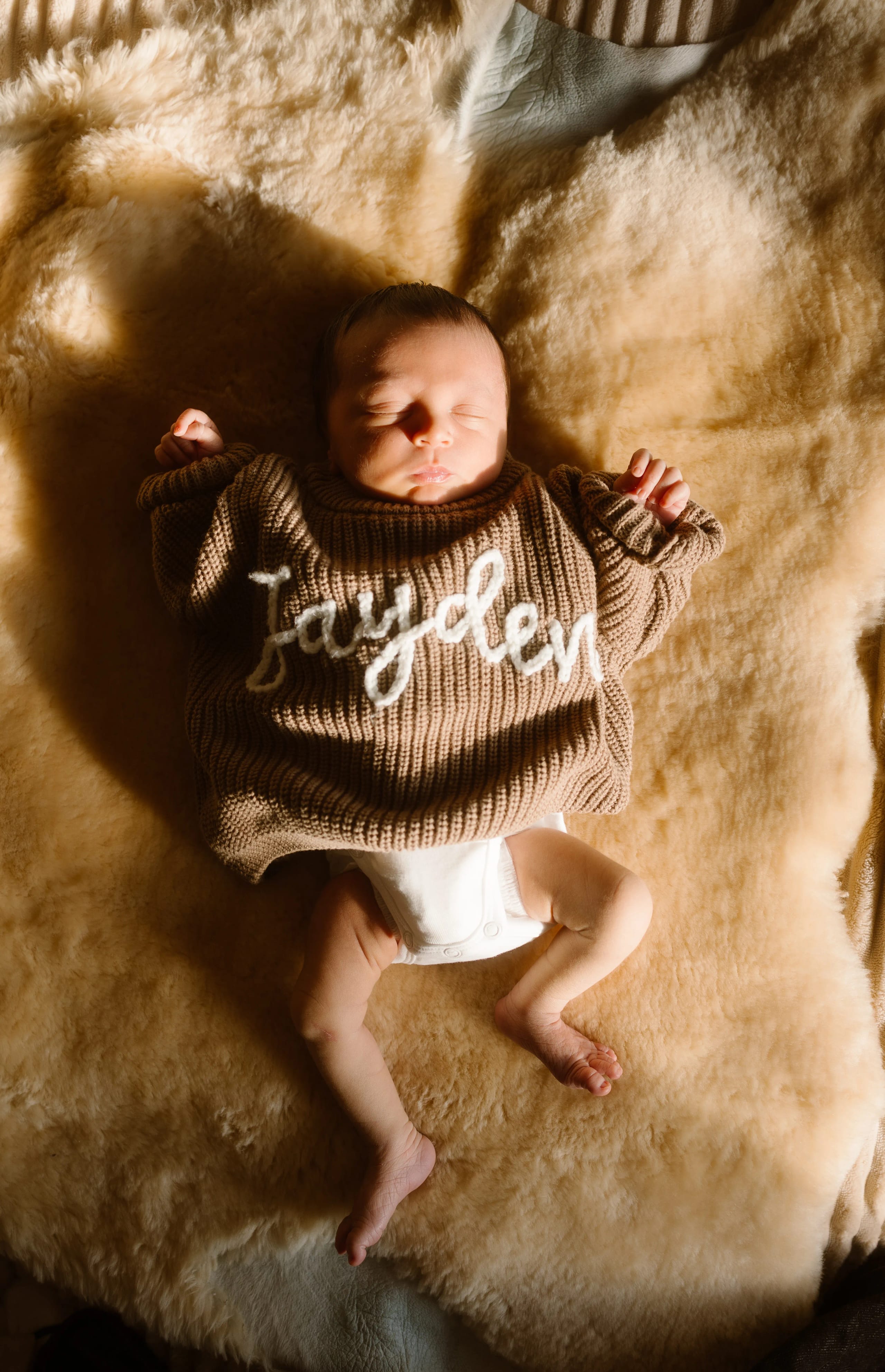 Een schattige baby ligt ontspannen op een bontachtige ondergrond, gekleed in een warme, gebreide trui met de naam "Jayden" erop. De baby heeft zijn ogen gesloten en zijn armpjes zijn ontspannen naast zich uitgerold.