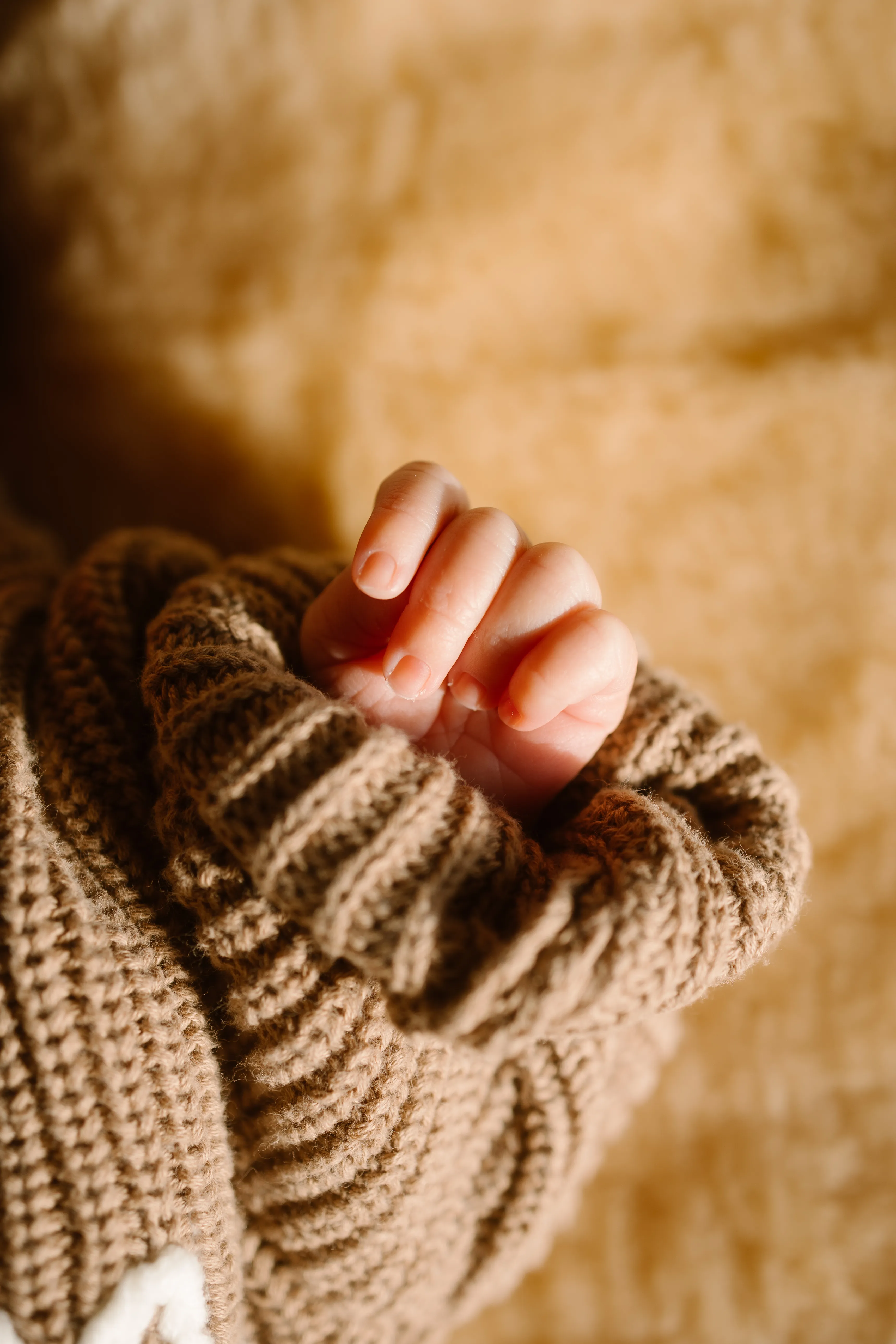 Een kleine hand van een baby die zich knus in een gebreide tan kleurige trui nestelt, omringd door een zachte, beige achtergrond.