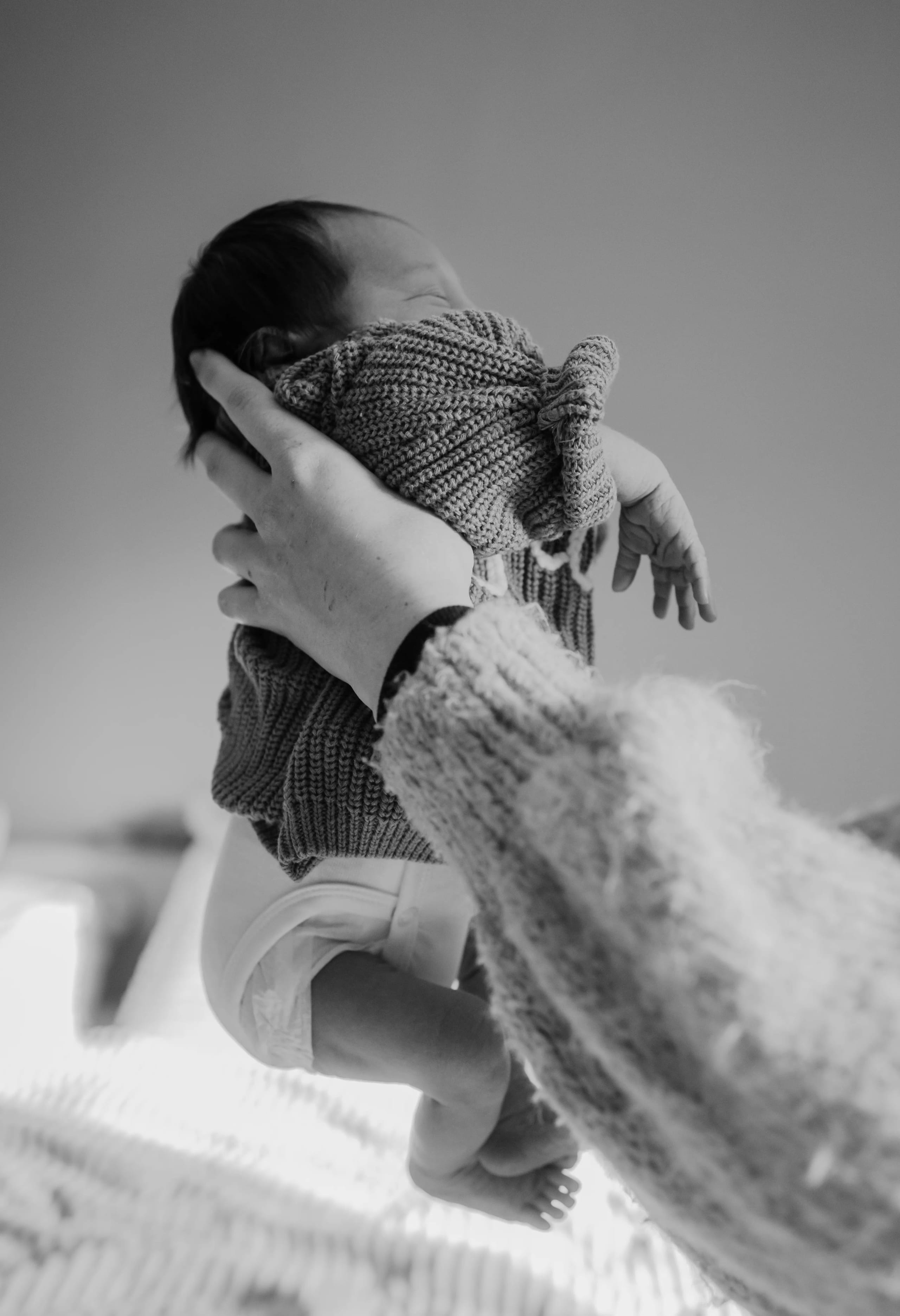 Een liefdevolle hand houdt een pasgeboren baby vast, gekleed in een gebreid jasje, terwijl the baby met een lichte glimlach naar voren kijkt, op een zachte ondergrond in een sereen interieur.