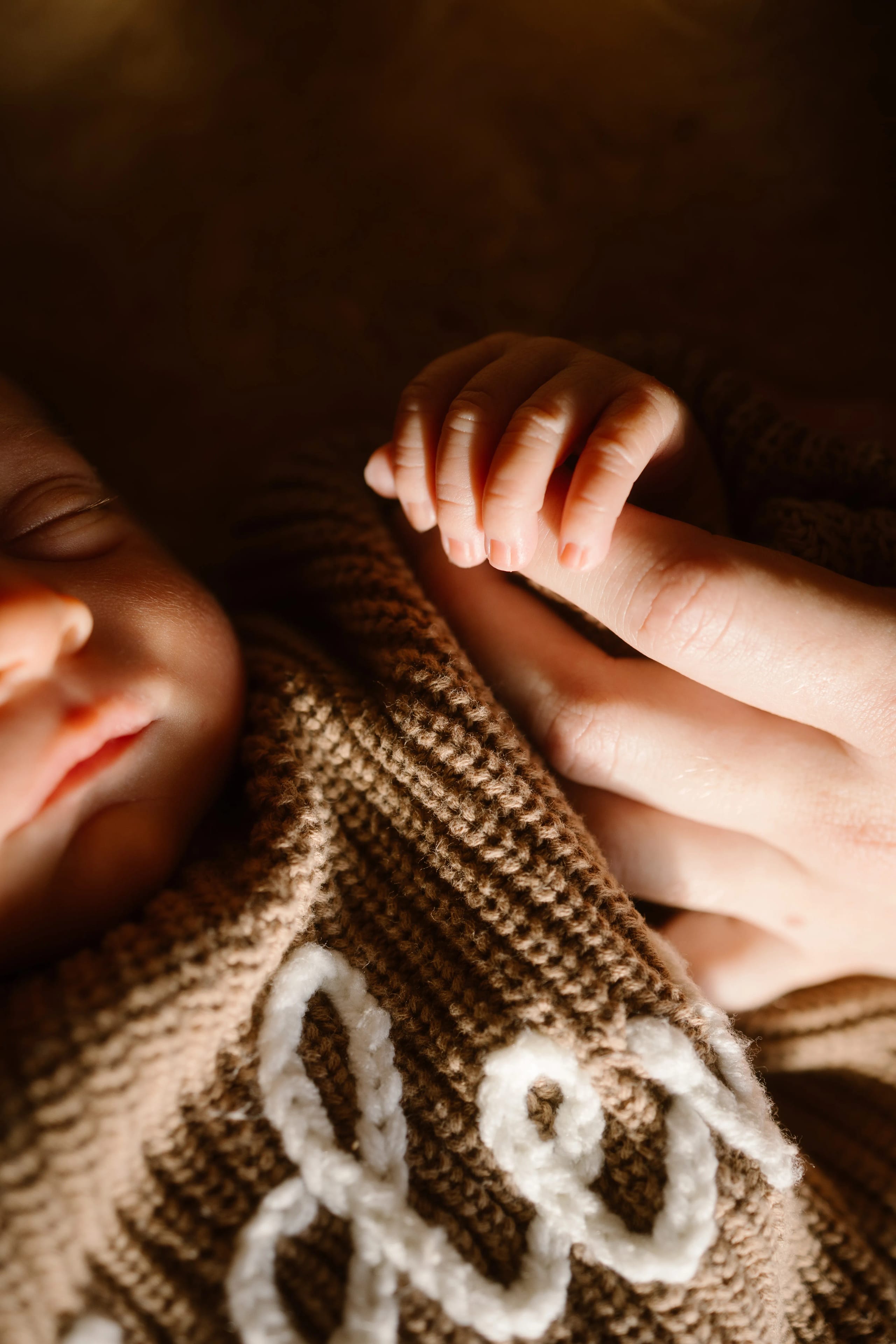 Een pasgeboren baby gewikkeld in een zachte, bruine deken, terwijl een volwassen hand voorzichtig de kleine hand van de baby vasthoudt, wat een intiem moment van verbondenheid en liefde uitstraalt.