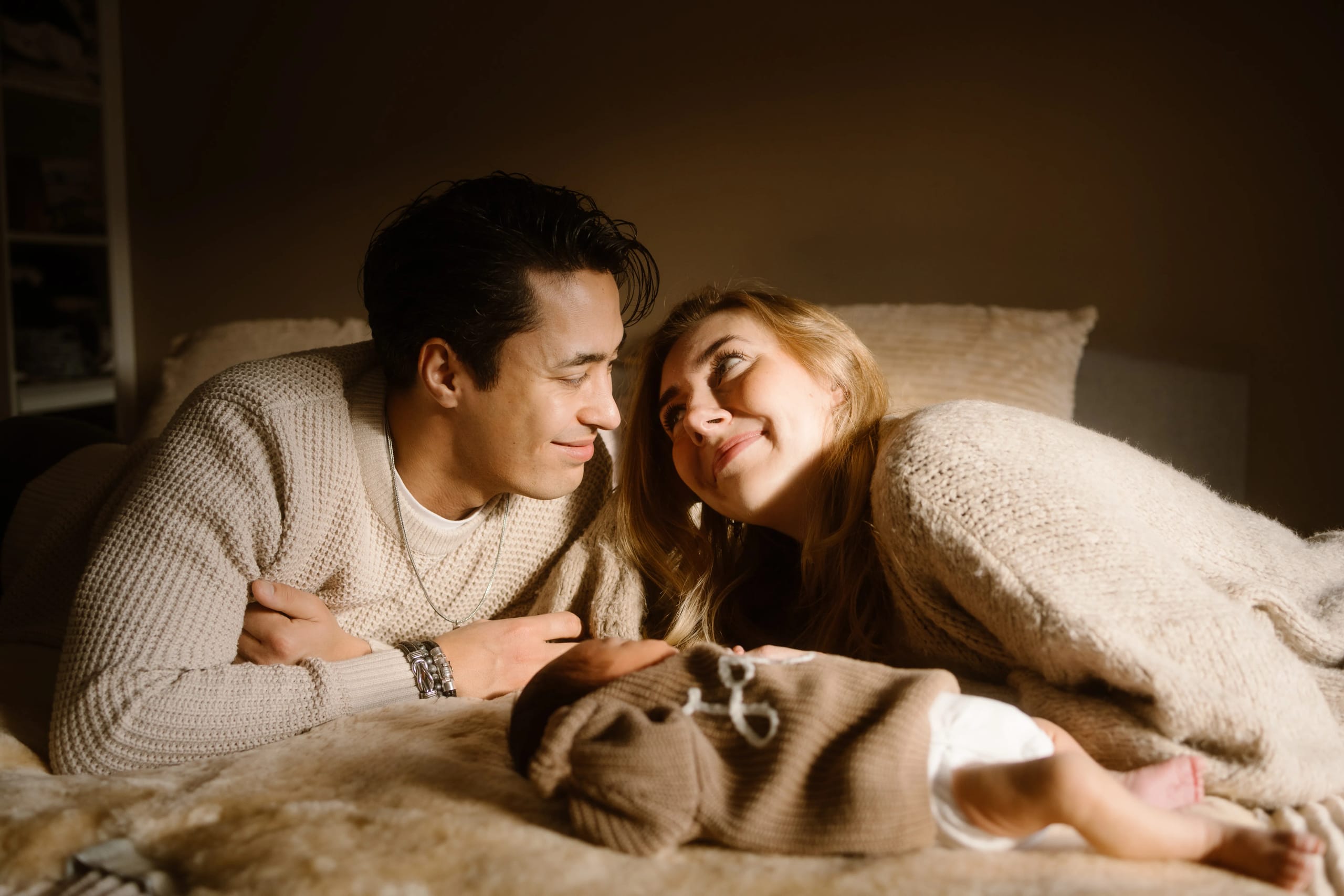 Gelukkige ouders liggen naast hun pasgeboren baby, knapperig in een warme, neutrale omgeving. Ze kijken elkaar liefdevol aan, omringd door een zachte sfeer die de intimiteit van het gezinsleven benadrukt.
