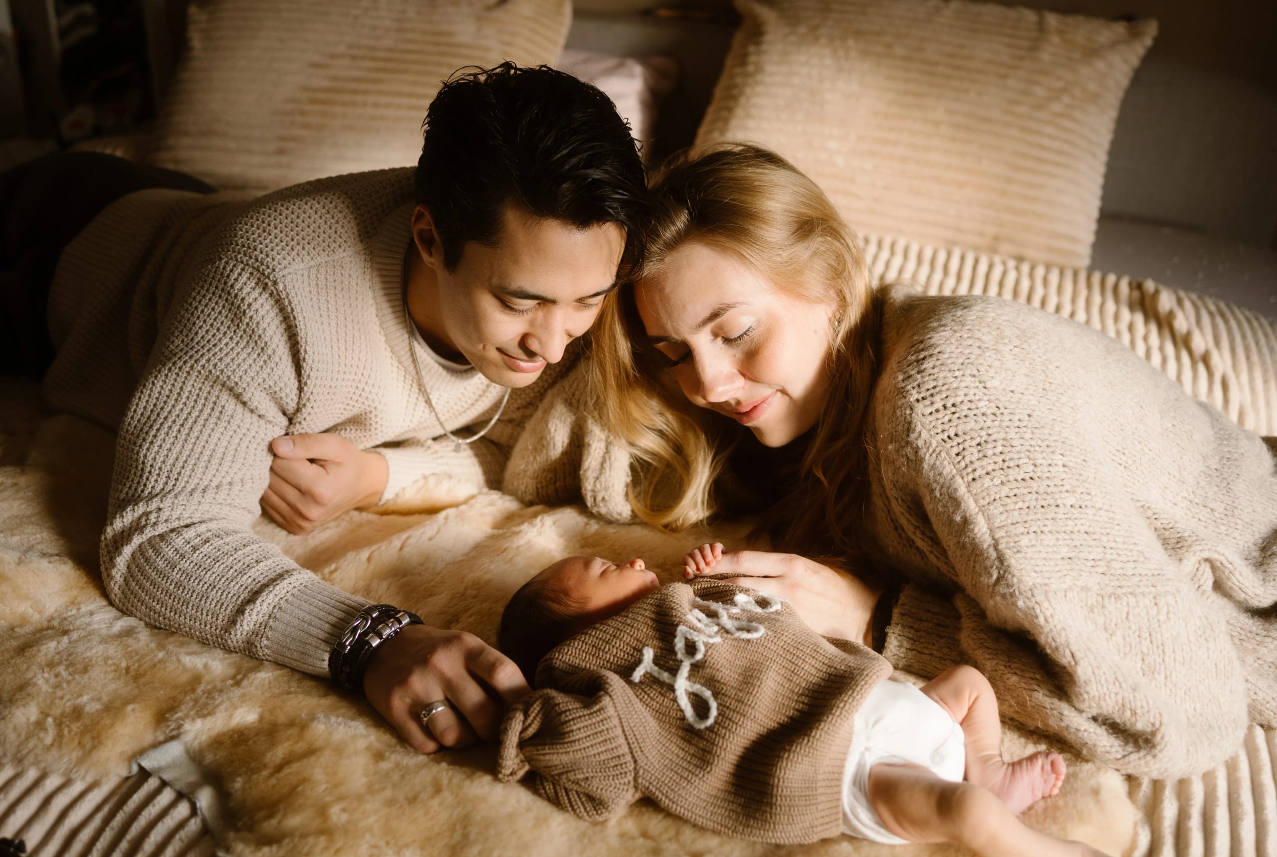 Een blije jonge familie ligt samen op een zachte deken, waarbij de ouders liefdevol naar hun pasgeboren baby kijken, gekleed in een schattige trui. De warme, serene sfeer wordt versterkt door het zachte licht dat de intieme momenten van samen zijn en het ouderschap vastlegt.