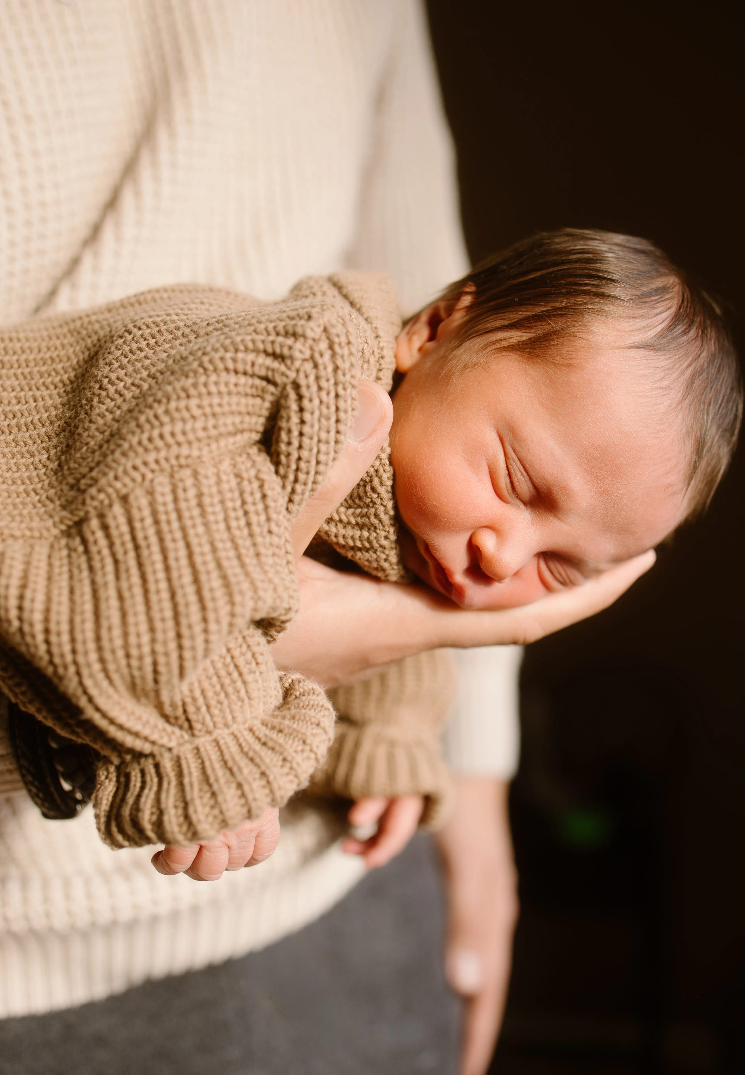 Een pasgeboren baby ligt vredig in de hand van een volwassene, gekleed in een warme, beige trui, met zachte, natuurlijke verlichting die de intieme sfeer benadrukt.