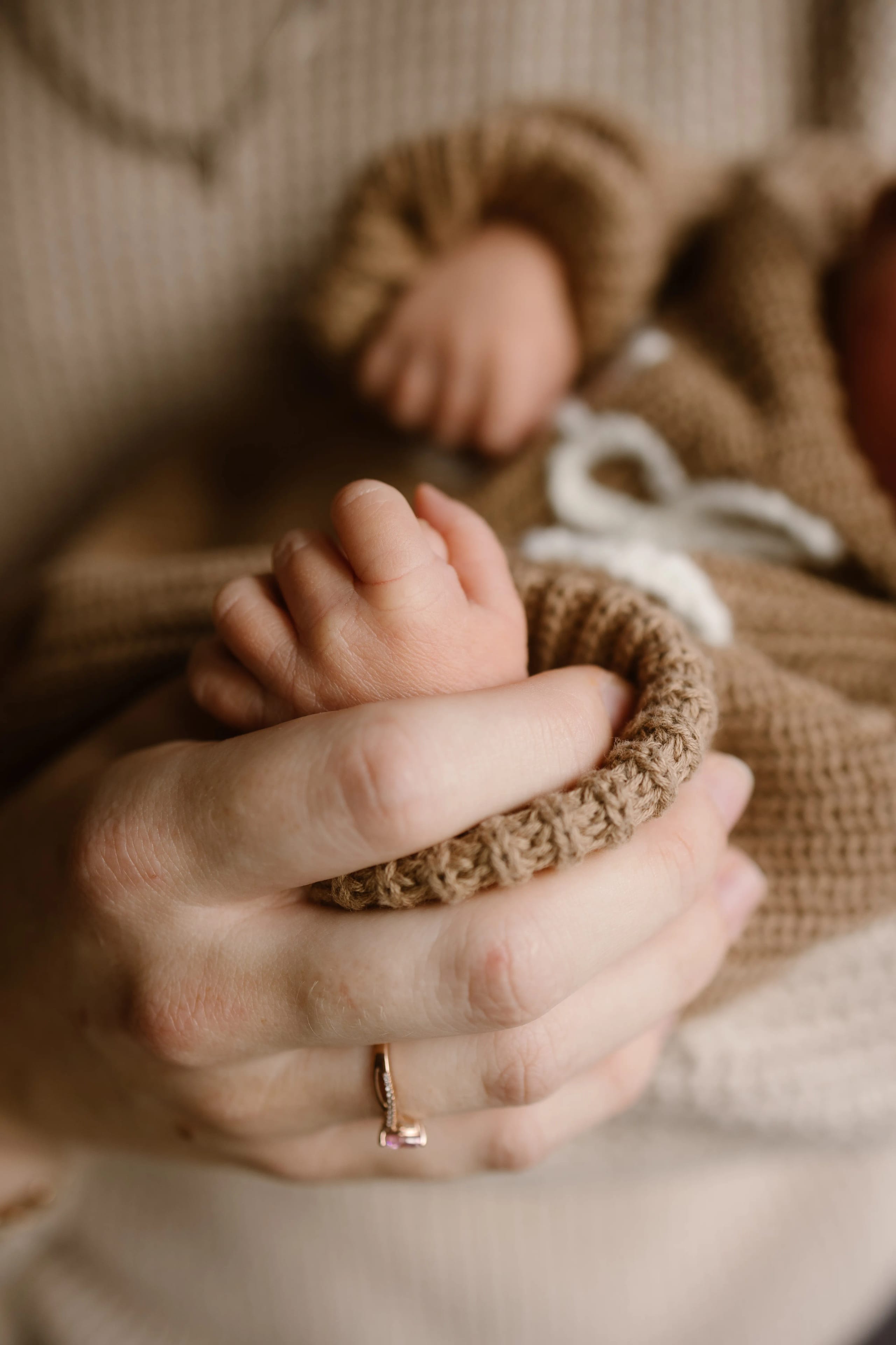Een menselijke hand houdt voorzichtig de kleine voet van een pasgeboren baby vast, gekleed in een schattige, bruingekleurde kleding. De focus ligt op de tedere aanraking en de warme verbinding tussen ouder en kind.