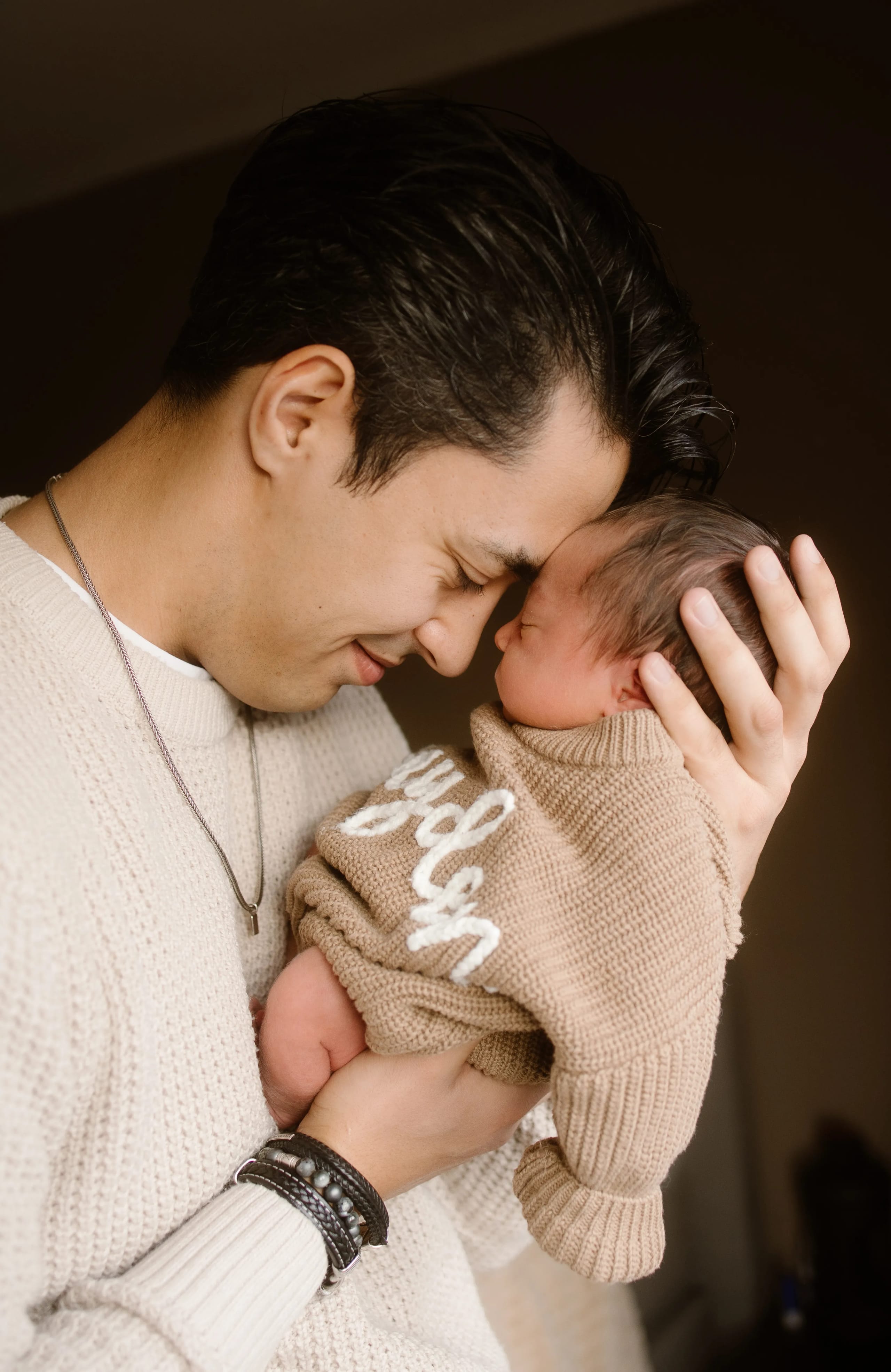 Een liefdevolle vader die zijn newborn met een beige trui vasthoudt, terwijl ze elkaar diep in de ogen kijken en hun voorhoofden bijna elkaar raken, wat een intieme en warme moment van verbinding weergeeft.