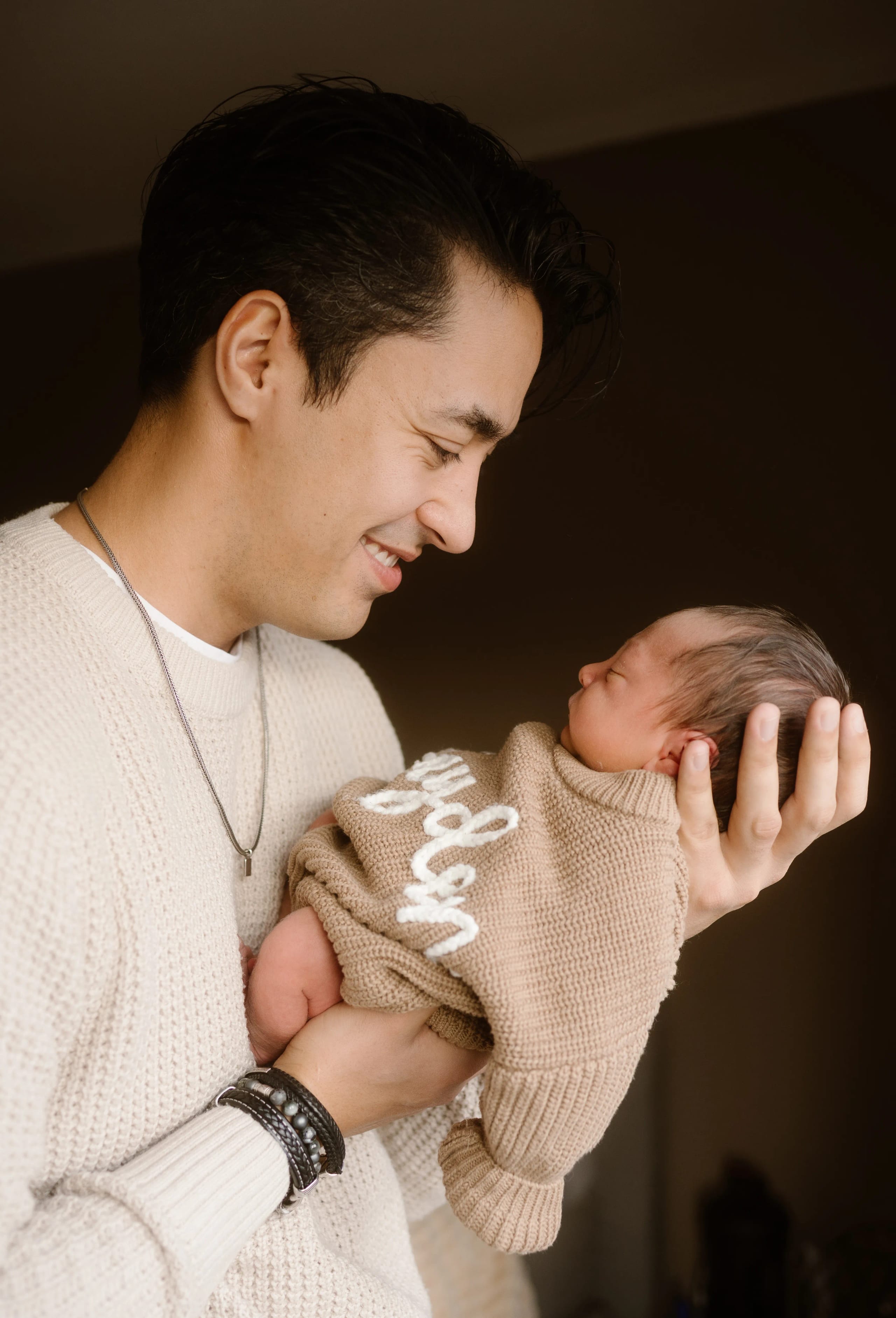 Een jonge vader kijkt liefdevol naar zijn pasgeboren baby, die in zijn arm gewikkeld is in een zachte, beige trui met de tekst "Jayden" erop. De warme en intieme sfeer benadrukt de speciale band tussen ouder en kind.
