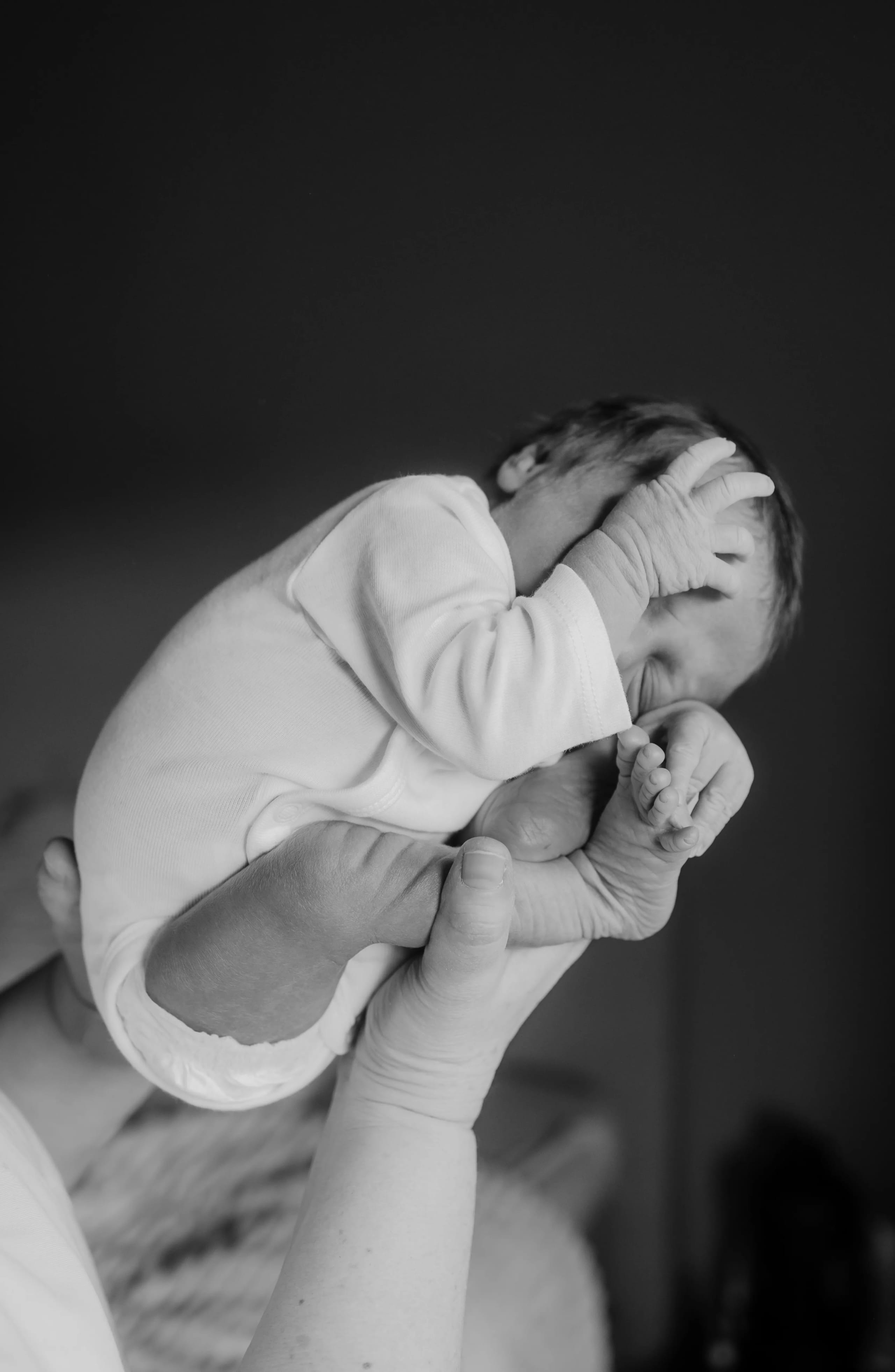 Een pasgeboren baby wordt liefdevol vastgehouden door een hand, terwijl hij of zij in een schattige houding met hun voeten naar boven ligt, met een zachte achtergrond die de intieme sfeer van het moment benadrukt.