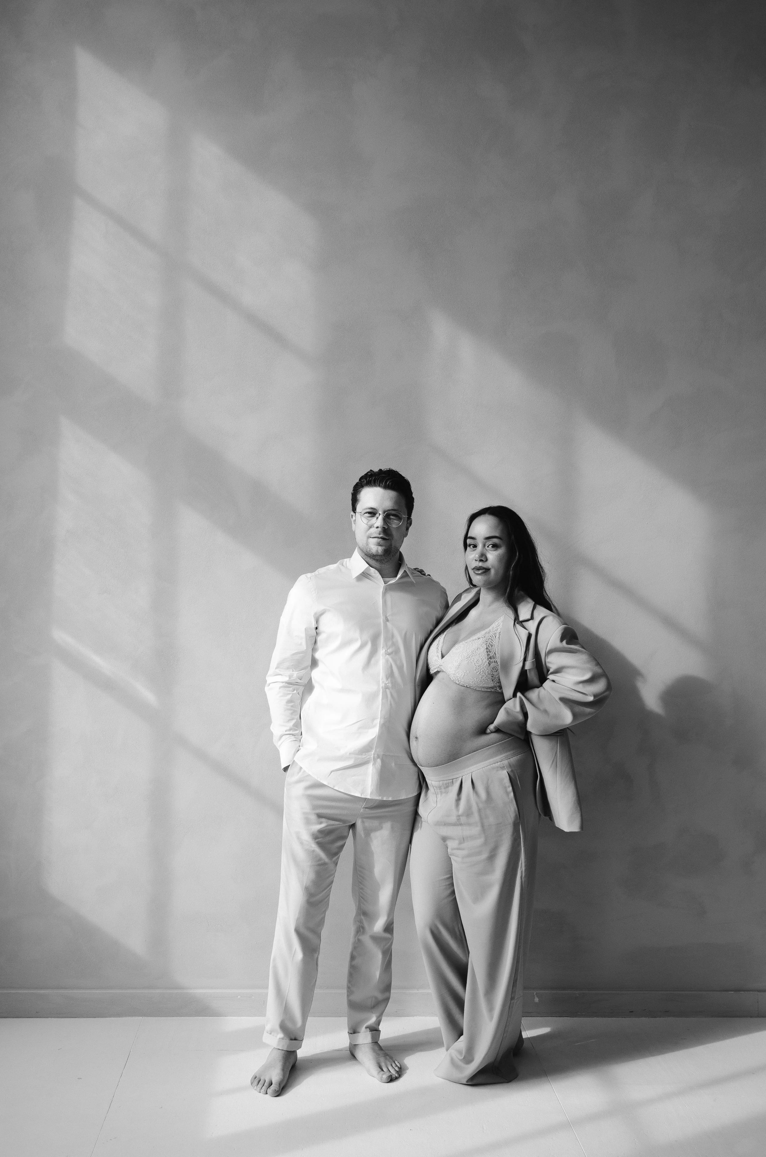Een aanstaande moeder in een stijlvolle outfit poseert trots naast haar partner, terwijl zij elkaar liefdevol aankijken in een natuurlijke lichtinval, waardoor schaduwen op de muur worden gecreëerd.