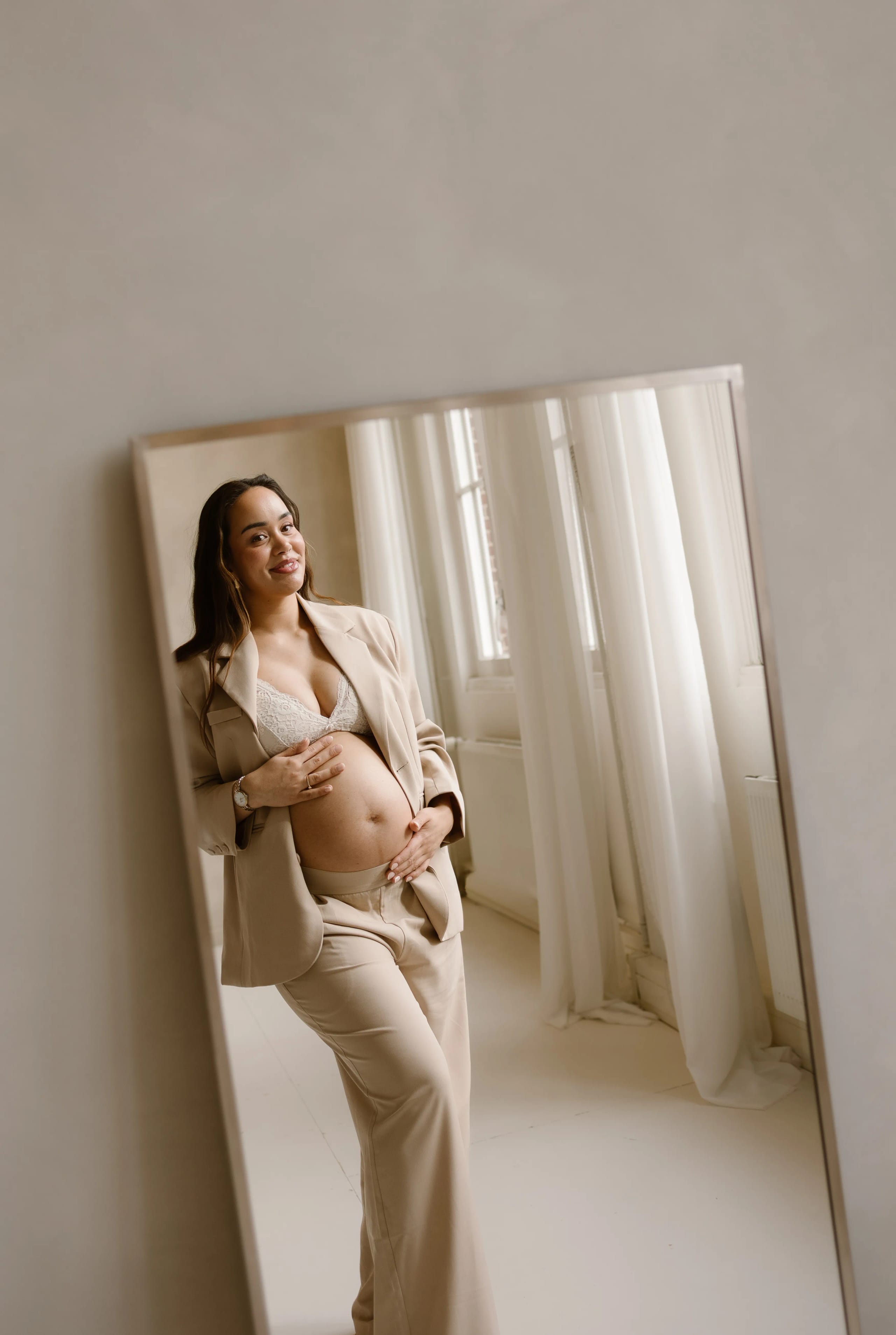 Een zwangere vrouw in een stijlvolle beige kostuum, glimlachend terwijl ze haar buik aanraakt, reflecteert in een grote spiegel, omringd door natuurlijke lichtinval en witte gordijnen.