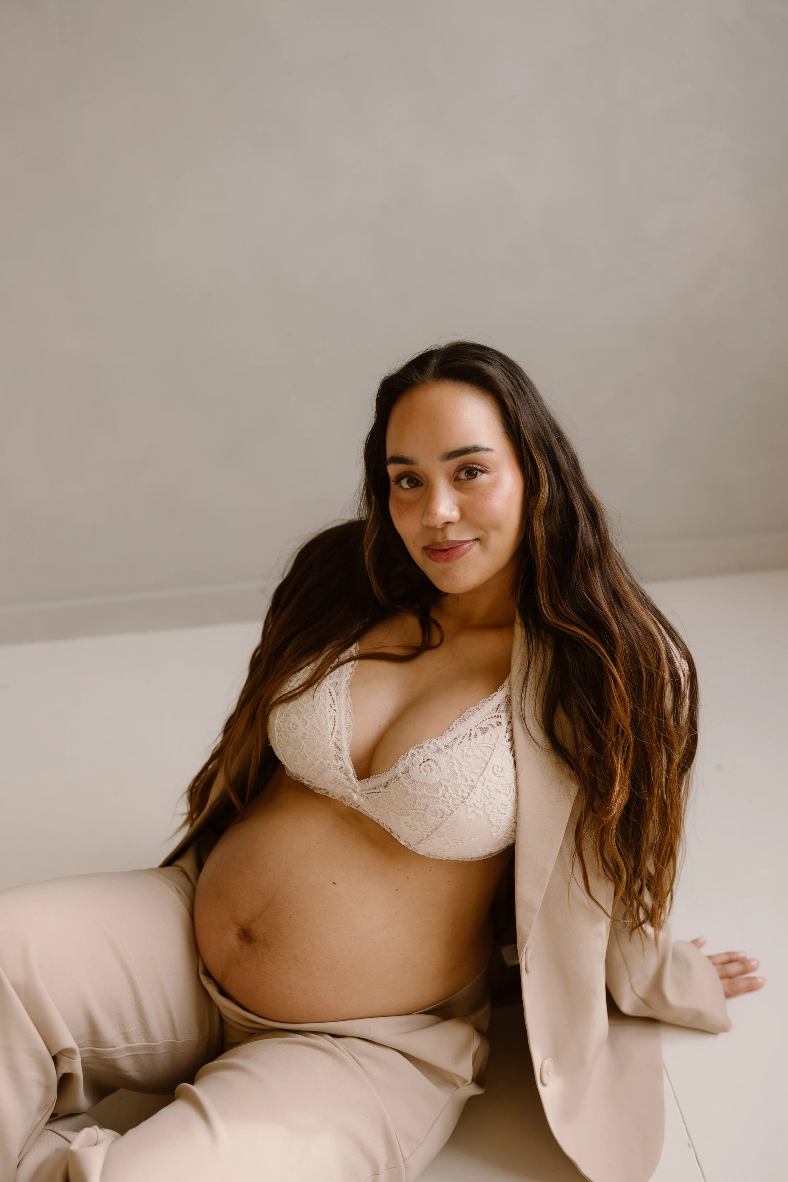 Zwangere vrouw zittend op de vloer in een stijlvolle beige broekpak, met een kanten bh, straalt zelfvertrouwen uit terwijl ze haar blik naar de camera richt. Haar lange, golvende haren en blijde uitdrukking benadrukken de schoonheid van de zwangerschap.