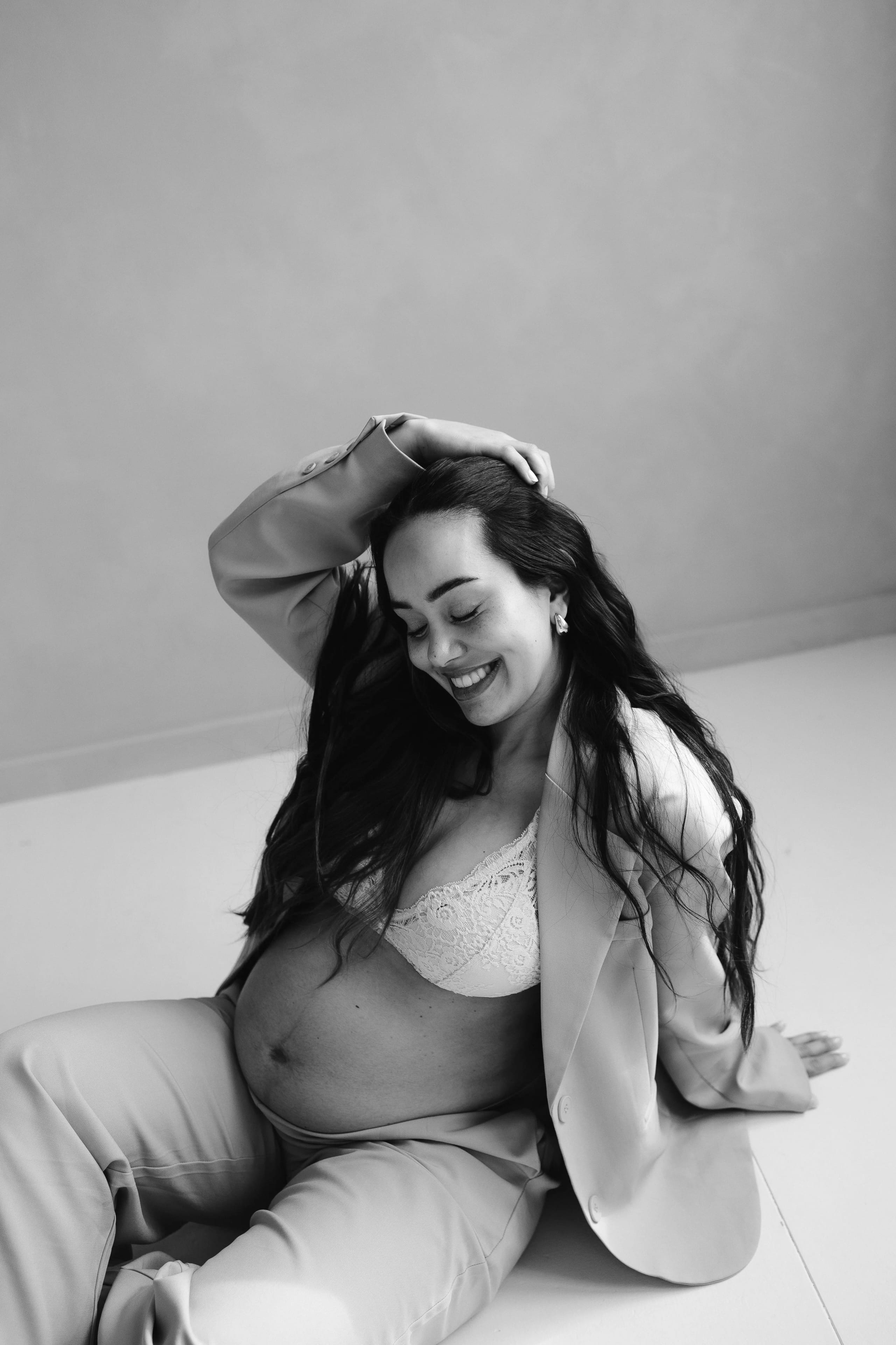 Zwangere vrouw met een glimlach, gekleed in een stijlvolle broek en blazer, zit ontspannen op de grond met een hand in haar haar en toont haar mooie, volle buik, tegen een neutrale achtergrond.