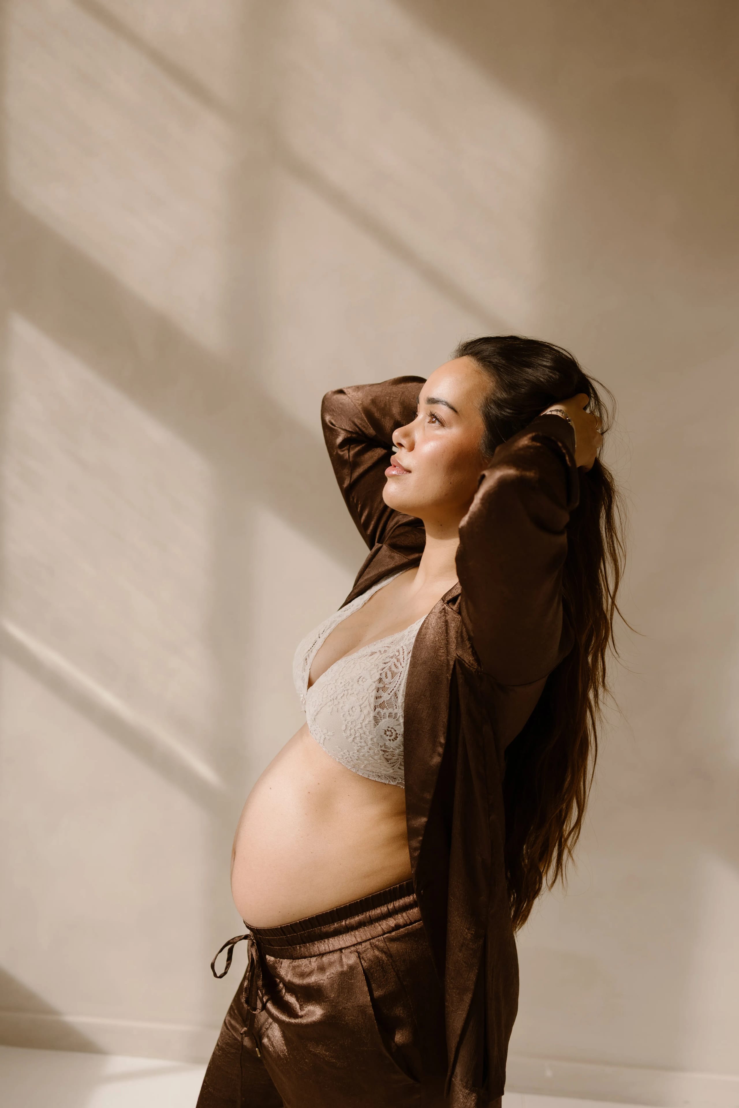 Een zwangere vrouw poseert elegant in een lichte ruimte, met haar handen in haar haar en een zelfverzekerde uitstraling, gekleed in een delicate beha en een bruine satijnen set. De zachte verlichting creëert een serene sfeer en benadrukt haar babybuik.