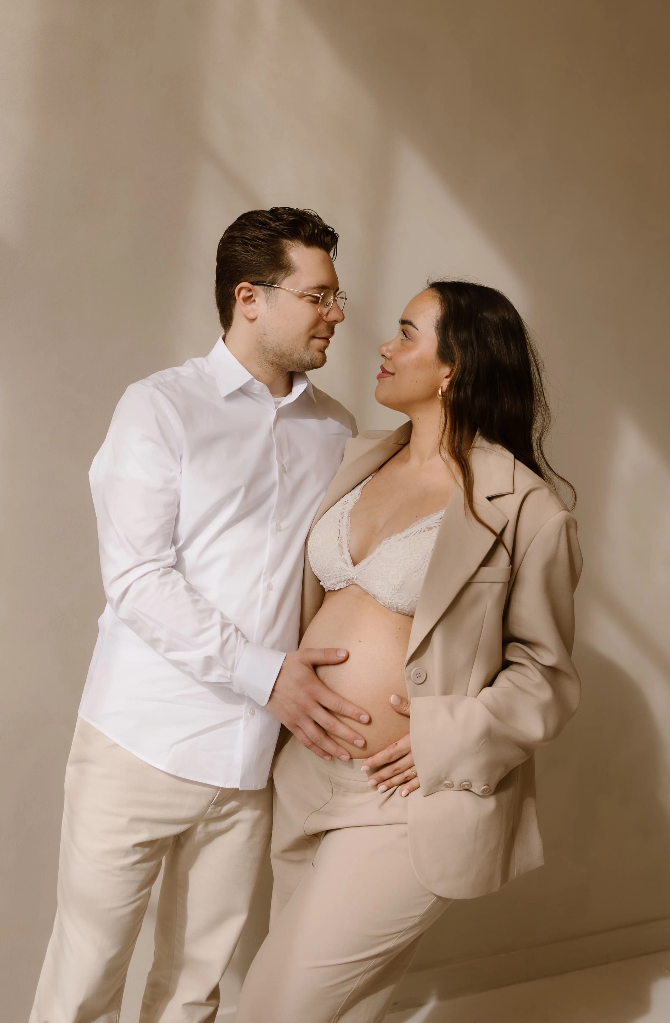 Een verliefd stel staat dicht bij elkaar in een lichte, neutrale omgeving, waarbij de man zijn hand op de zwangere buik van de vrouw legt. De vrouw draagt een elegante beige outfit en een lingerie-top, terwijl de man in een wit overhemd gekleed is.
