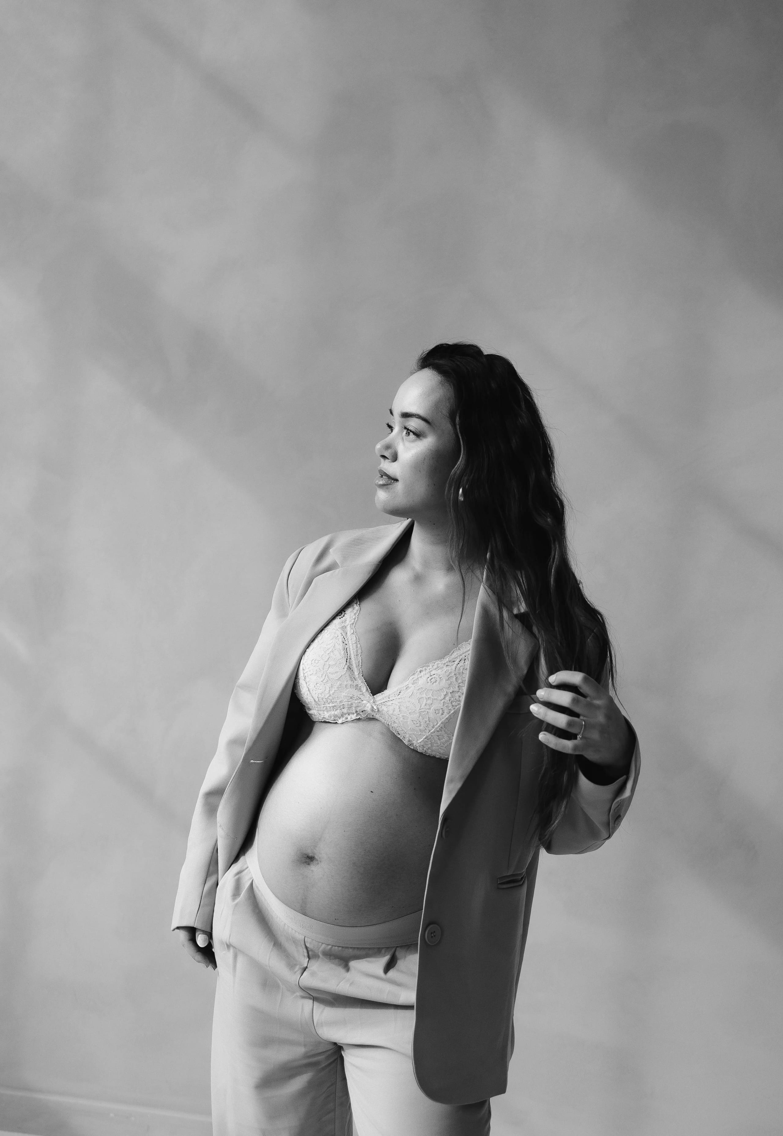 Een zwangere vrouw in een stijlvolle beige blazer en een glinsterende beha poseert met een zelfverzekerde uitstraling, met haar hand door haar lange haar terwijl ze naar de zijkant kijkt. De afbeelding is in zwart-wit, wat de elegantie en kracht van haar zwangerschap benadrukt.