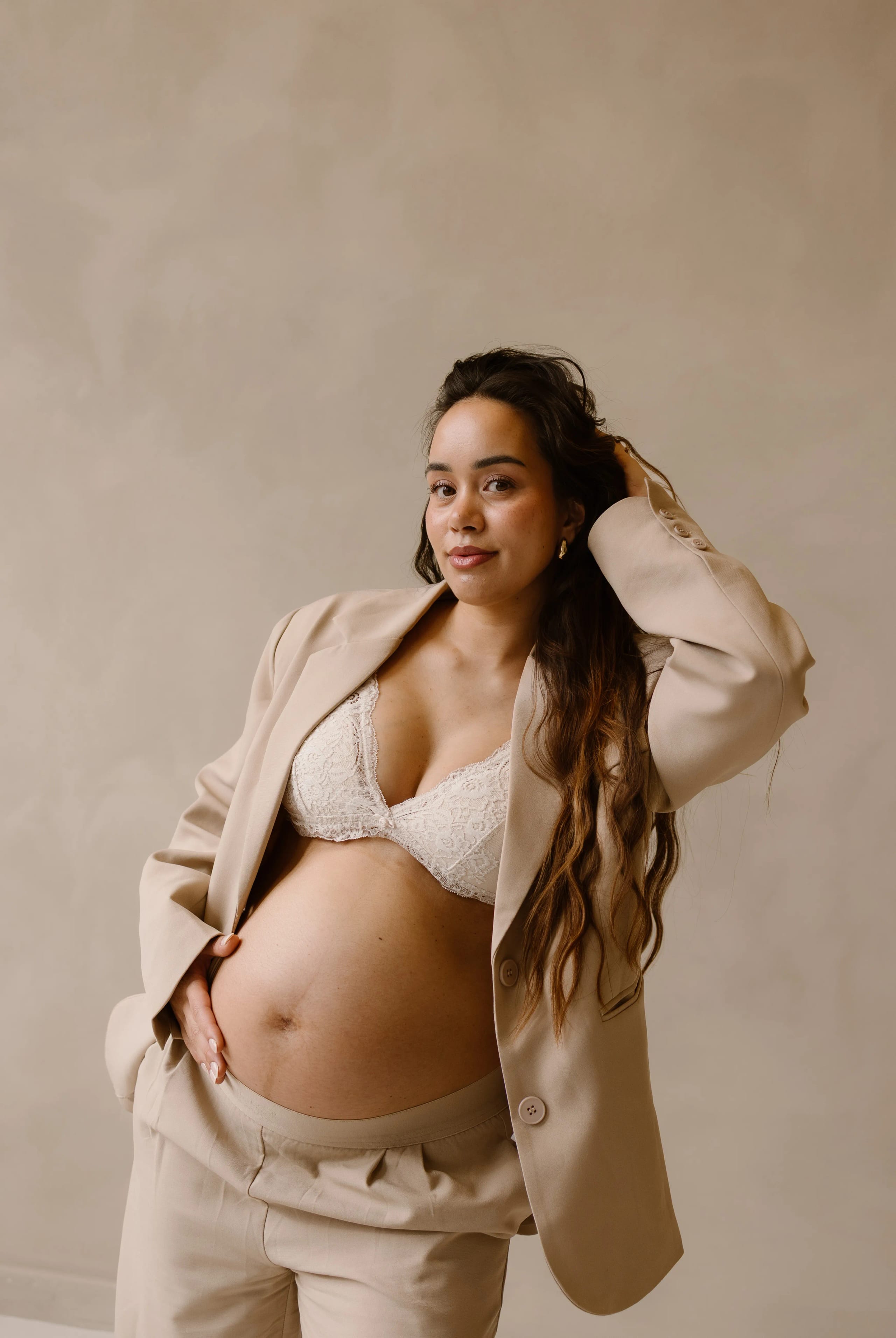 Zwangere vrouw in een stijlvolle beige blazer en broek poseert zelfverzekerd met haar hand op haar buik, voor een neutrale achtergrond.