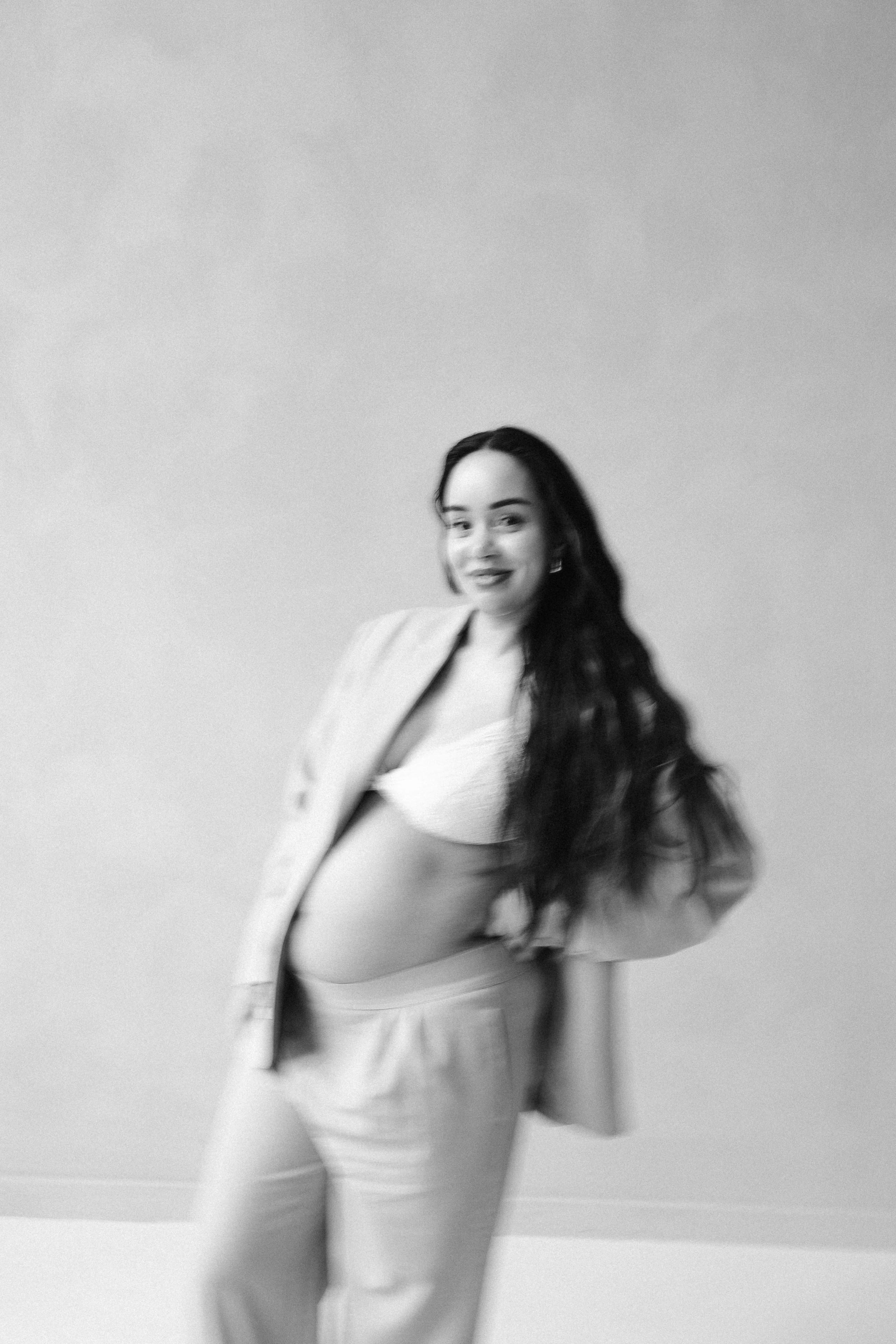 Een glimlachende zwangere vrouw poseert in een stijlvolle, casual outfit met een losse blazer en een crop top, terwijl ze haar buik trots toont tegen een neutrale achtergrond.