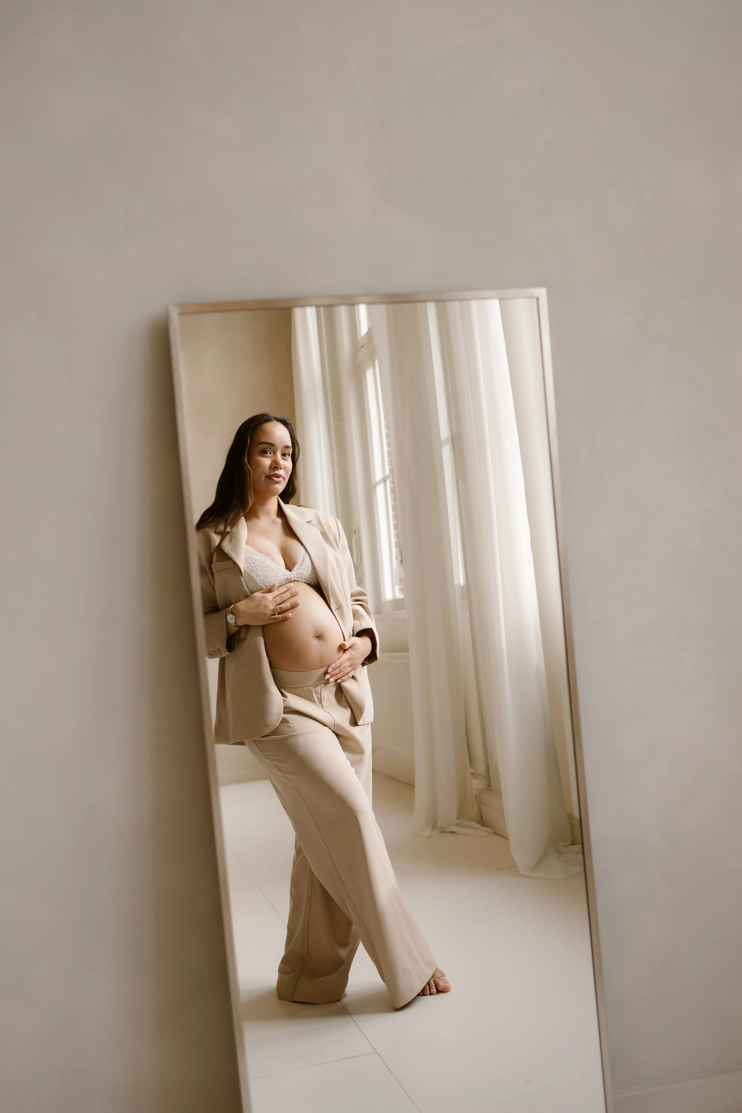 Zwangere vrouw in een stijlvolle combinatie van een beige blazer en broek, poseert zelfverzekerd voor een grote spiegel, terwijl ze haar buik aanraakt in een lichte, rustige omgeving met grote ramen en witte gordijnen.
