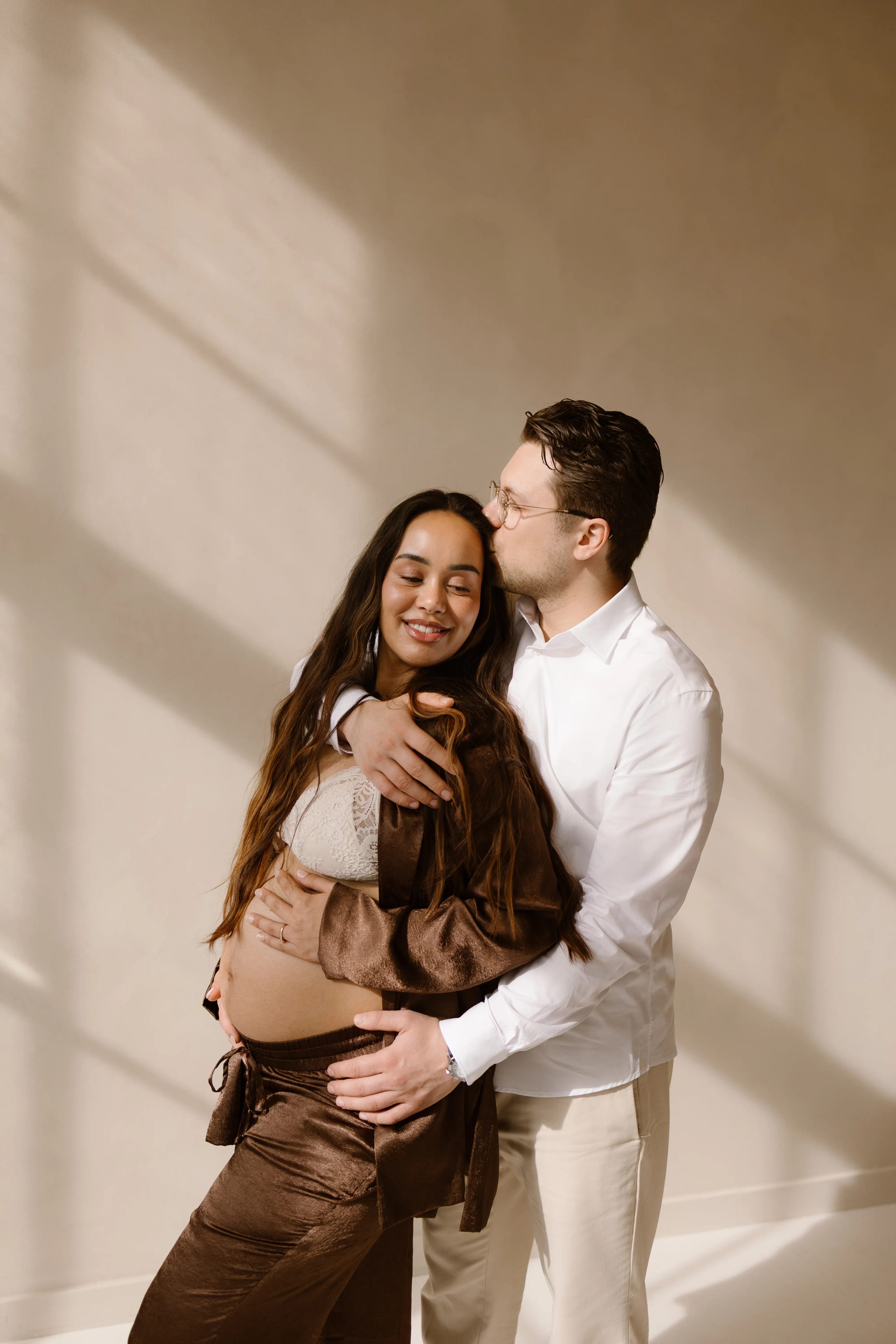 Een zwangere vrouw in een elegante bruine outfit staat knuffelend tegen haar partner, terwijl hij een tedere kus op haar hoofd geeft. Ze stralen liefde en blijdschap uit in een sfeervolle omgeving met zacht daglicht dat door het raam valt.