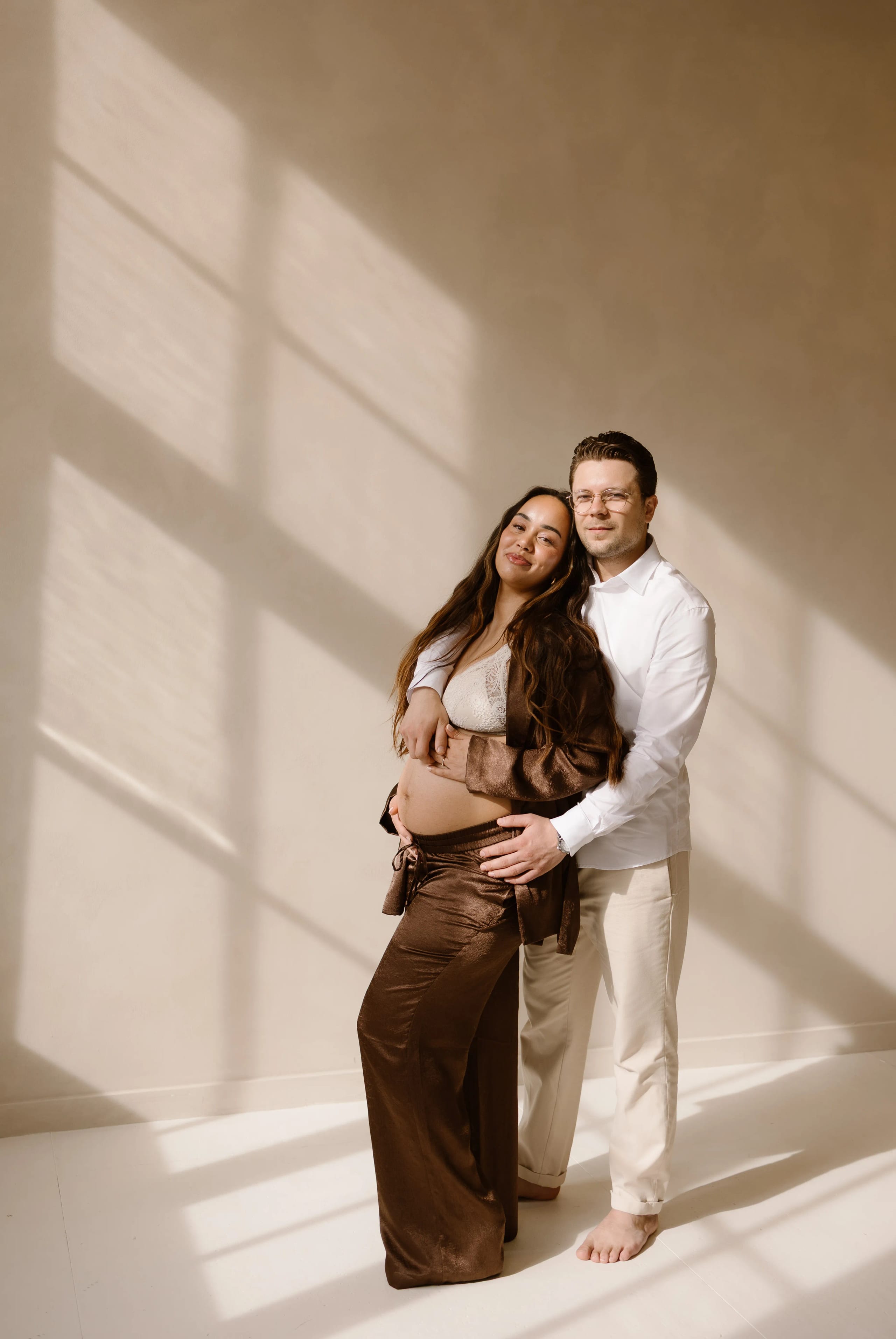Een zwangere vrouw in stijlvolle kleding poseert hoopvol terwijl ze haar buik omarmt, samen met haar partner die liefdevol achter haar staat, in een lichtovergoten ruimte met zachte schaduwen.