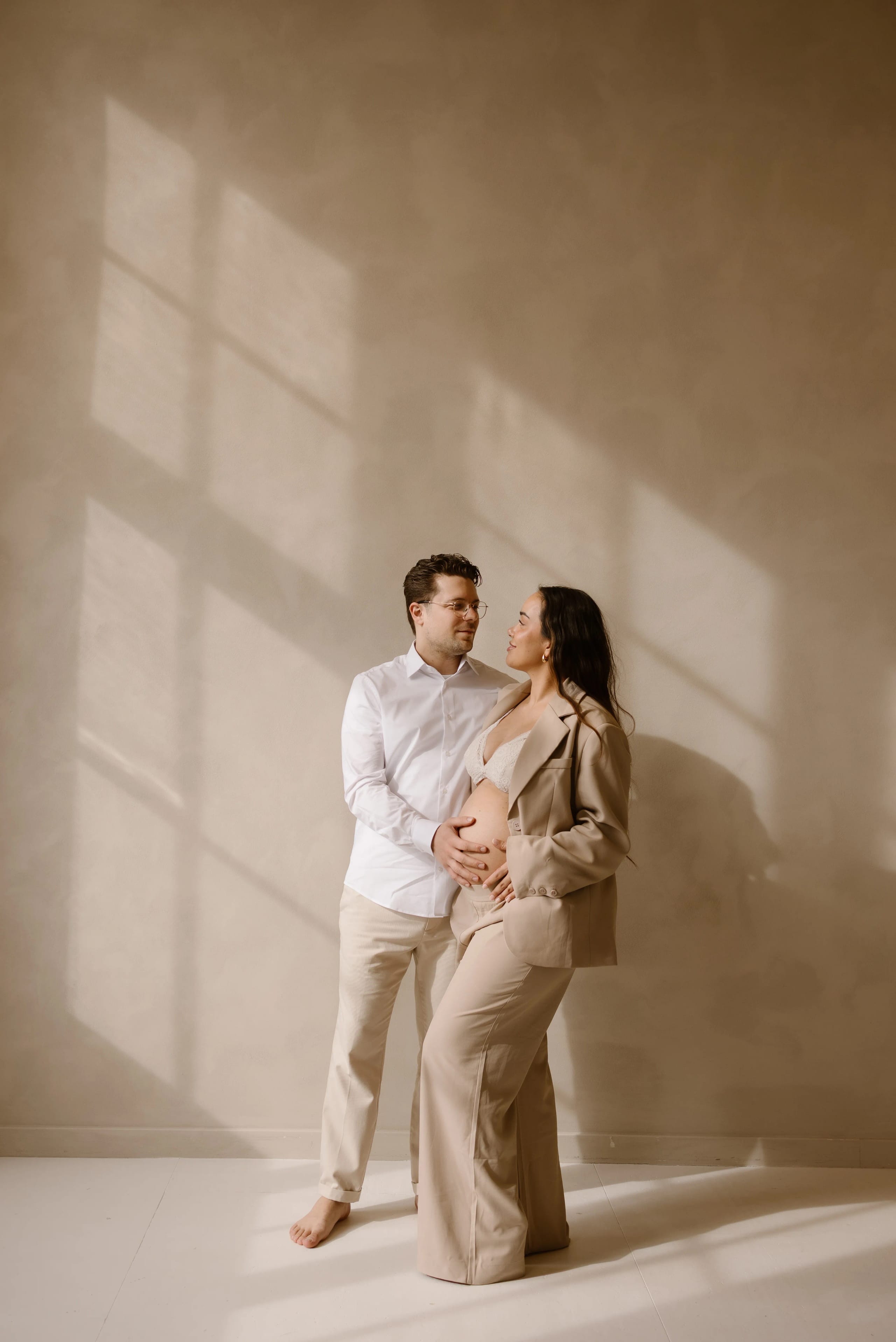 Een blije zwangere vrouw in een stijlvolle beige broek en blazer poseert liefdevol met haar partner, die een witte overhemd draagt, terwijl ze elkaar aankijken in een ruimte met prachtige lichtinval en zachte schaduwen op de muur.