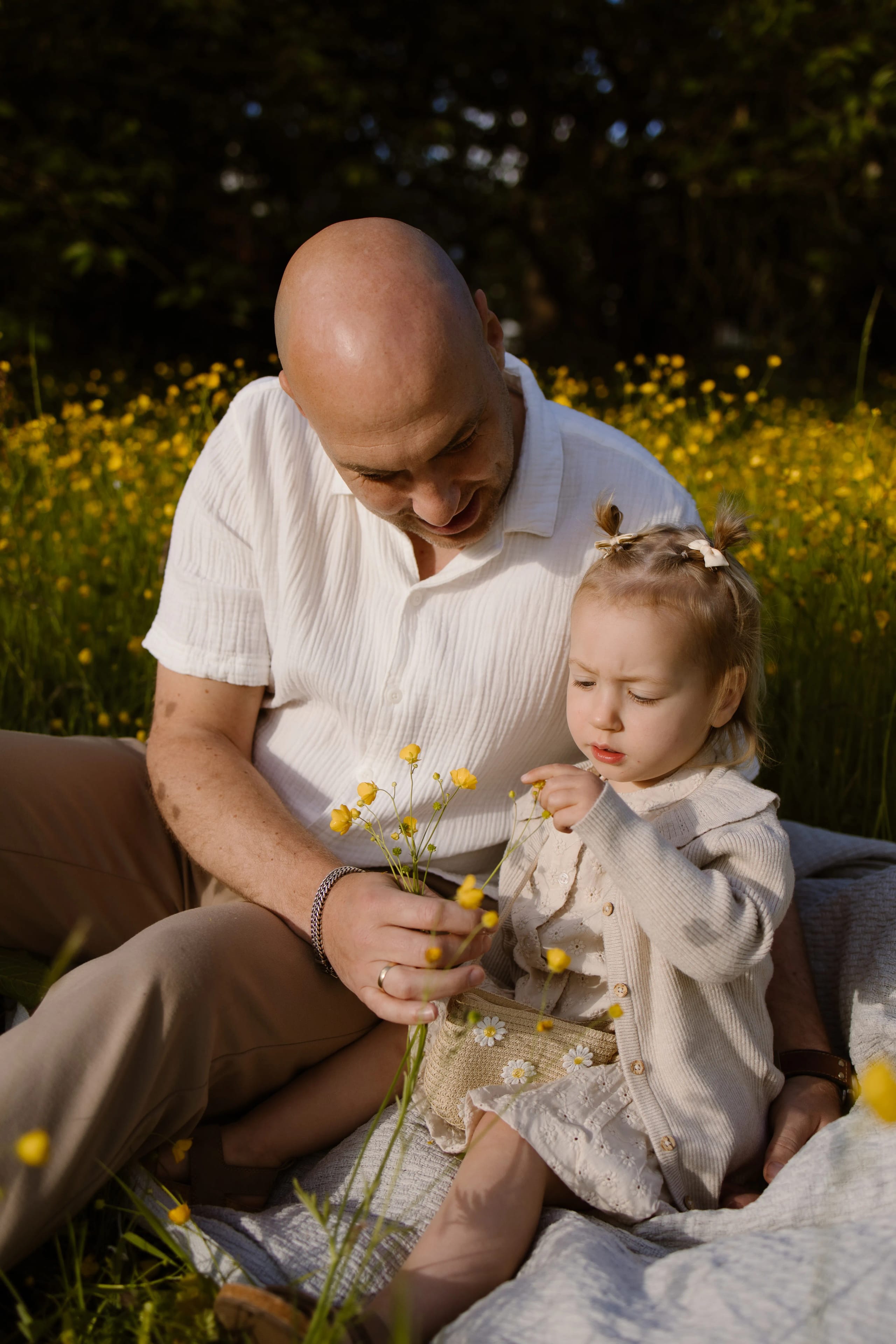 Vader en dochter zitten op een kleed in een bloeiende weiland, omringd door geel-florale boterbloemen. De vader lacht terwijl hij de bloemen in de hand van zijn kind toont, dat gefascineerd naar de bloemen kijkt en met zijn vinger wijst.