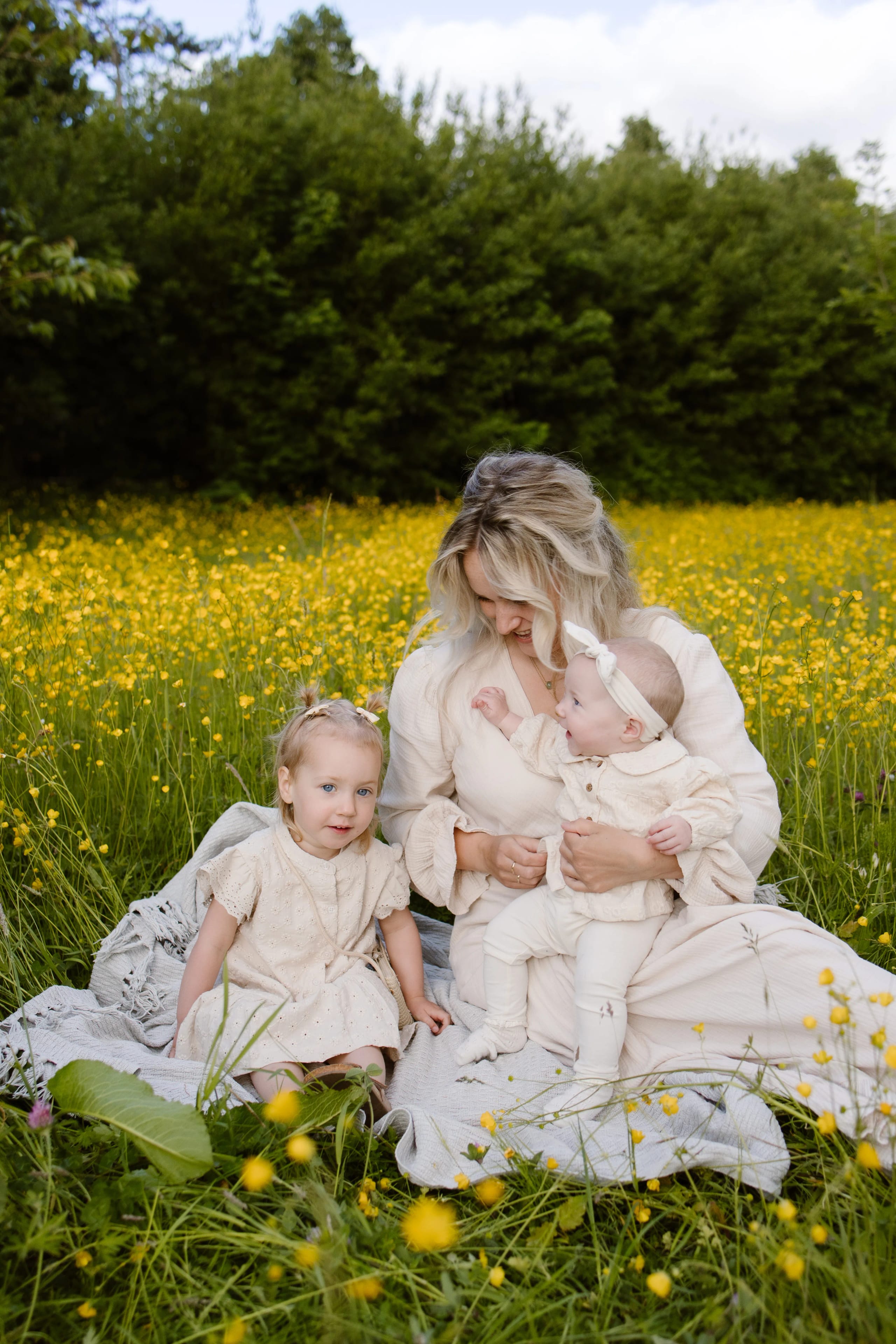 Een moeder zit op een deken in een veld vol gele boterbloemen, terwijl ze aandacht heeft voor haar twee jonge kinderen, die beide in lichte, schattige kleding zijn gekleed. De oudste dochter kijkt naar de camera met een vriendelijke glimlach, terwijl de jongste, geïntroduceerd door de moeder, nieuwsgierig om zich heen kijkt.
