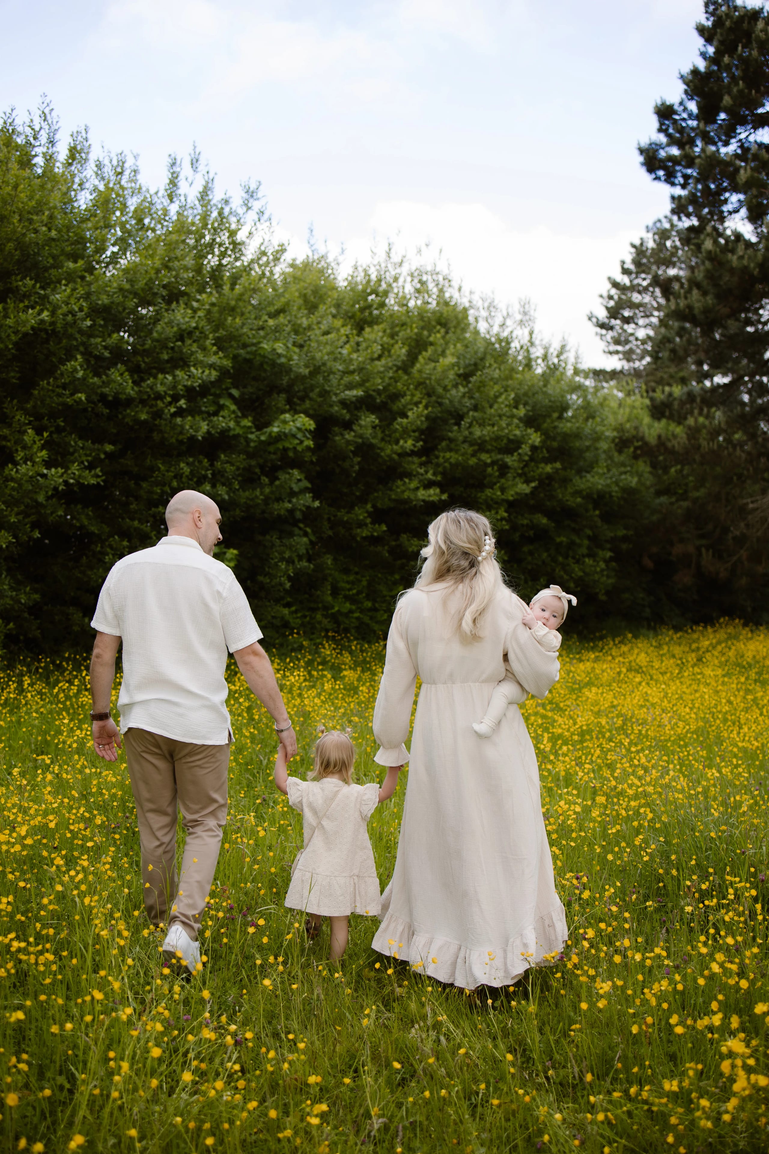 Een gezin wandelt hand in hand door een bloeiende weide vol gele bloemen, waarbij de vader en moeder hun kinderen begeleiden. De moeder houdt een baby vast terwijl de kleine met een lichtgekleurde jurk de hand van haar vader vasthoudt, onder een heldere lucht met enkele wolken.