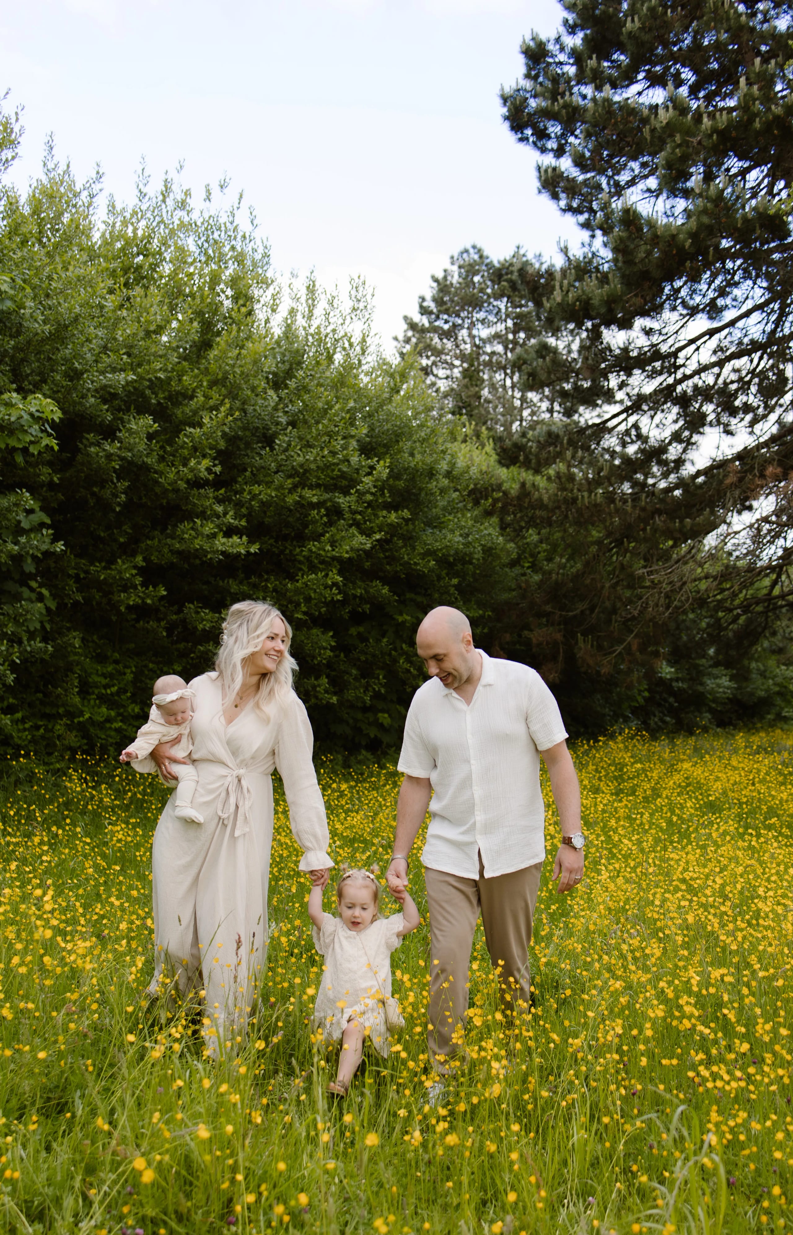 Een gezin wandelt samen door een bloeiende weide vol gele bloemen, waarbij de moeder een baby vasthoudt en de vader hand in hand loopt met hun peuter. Achter hen zijn weelderige bomen zichtbaar, wat een rustige en gelukkige buitenomgeving creëert.