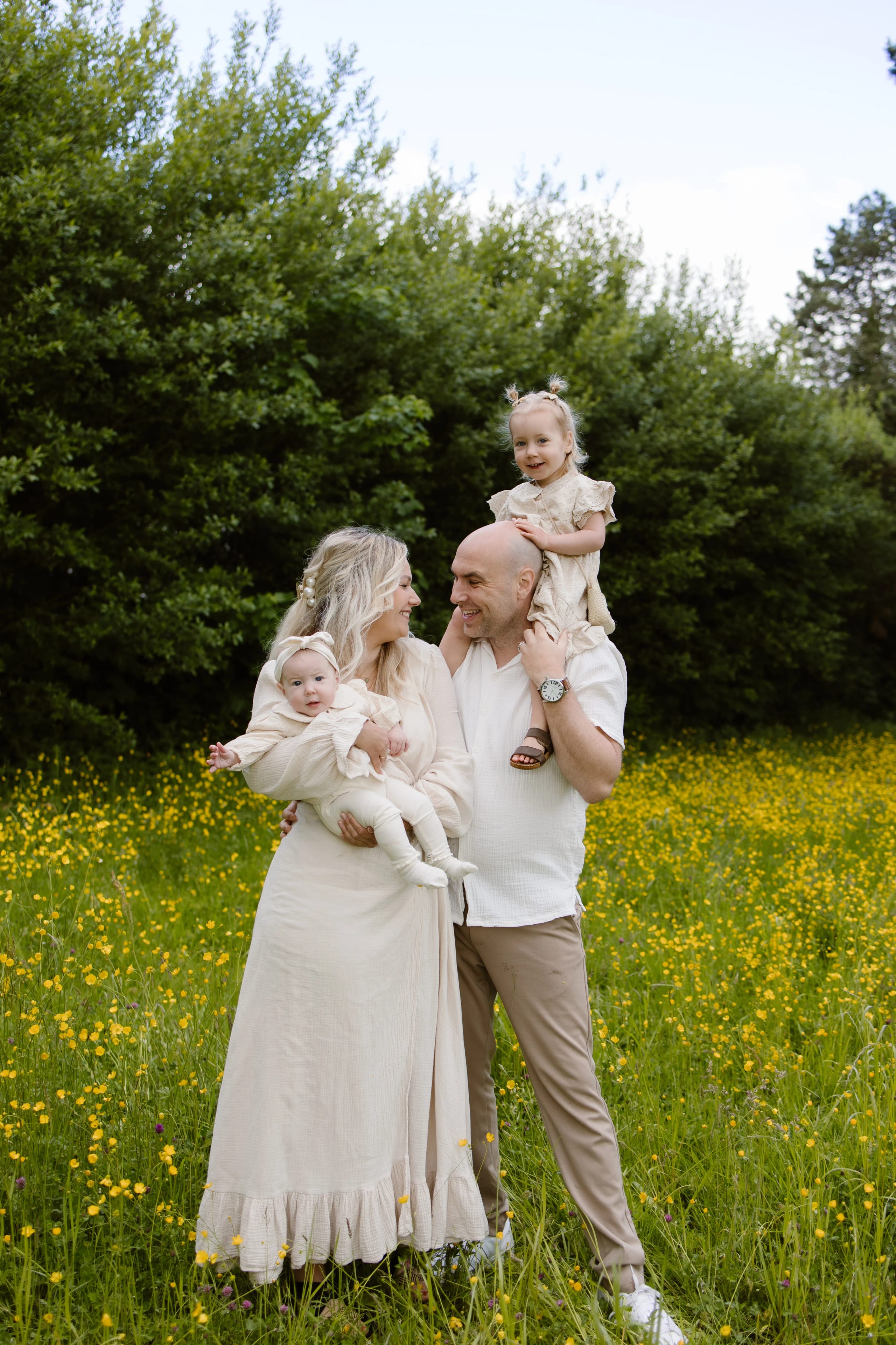 Een gelukkige familie staat in een bloeiende groene weide, met een moeder in een lange, lichte jurk die een baby vasthoudt, terwijl de vader met een blije peuter op zijn schouders staat. Achtergrond gevuld met groene struiken en kleurrijke bloemen benadrukt een gezellige, natuurlijke sfeer.