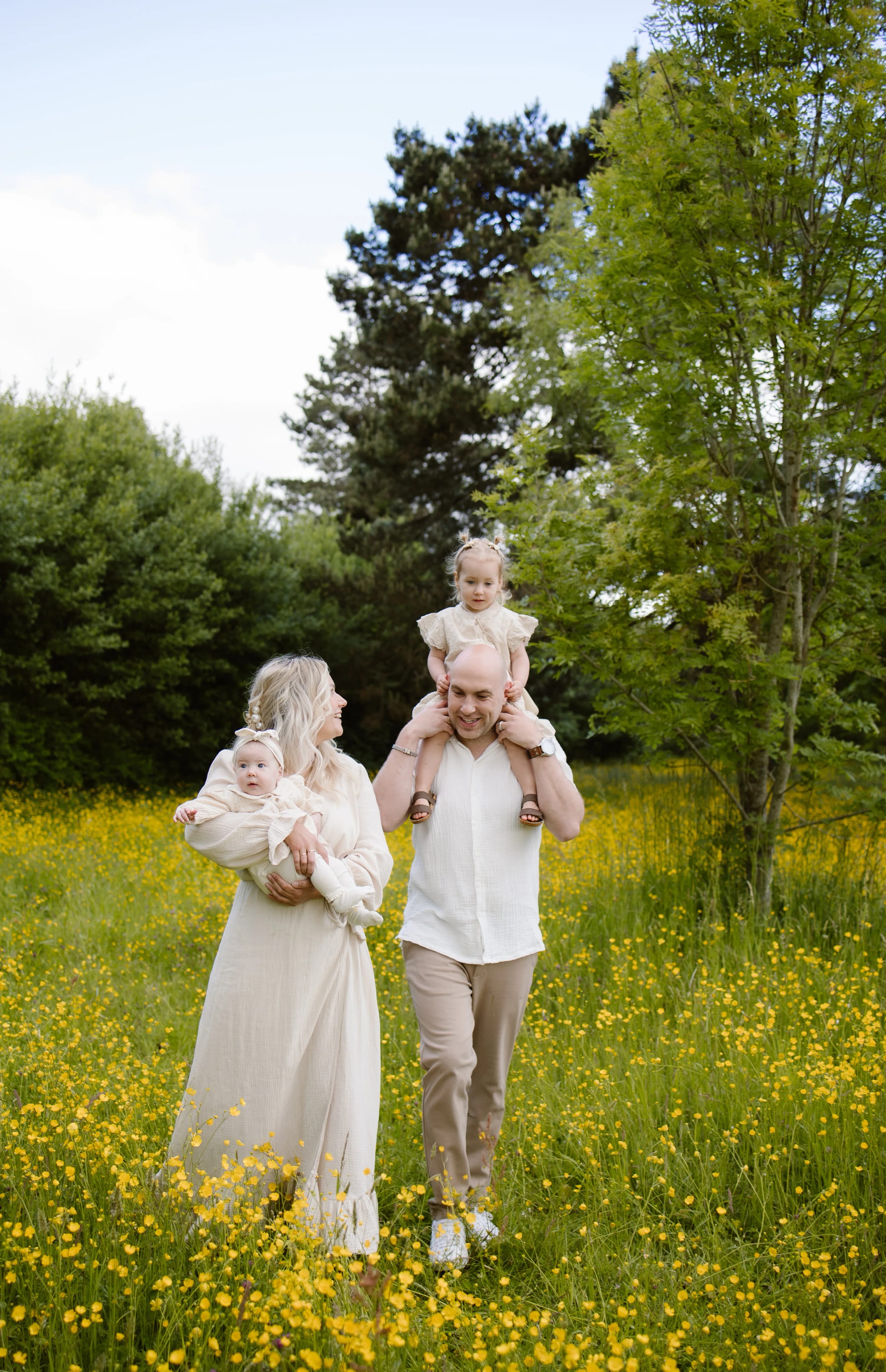 Een gelukkige familie wandelt door een zonovergoten bloemenveld, with a moeder die haar baby vasthoudt en een vader die zijn peuter op zijn schouders heeft. De achtergrond is omringd door groene bomen, wat een vrolijke en natuurlijke sfeer creëert.