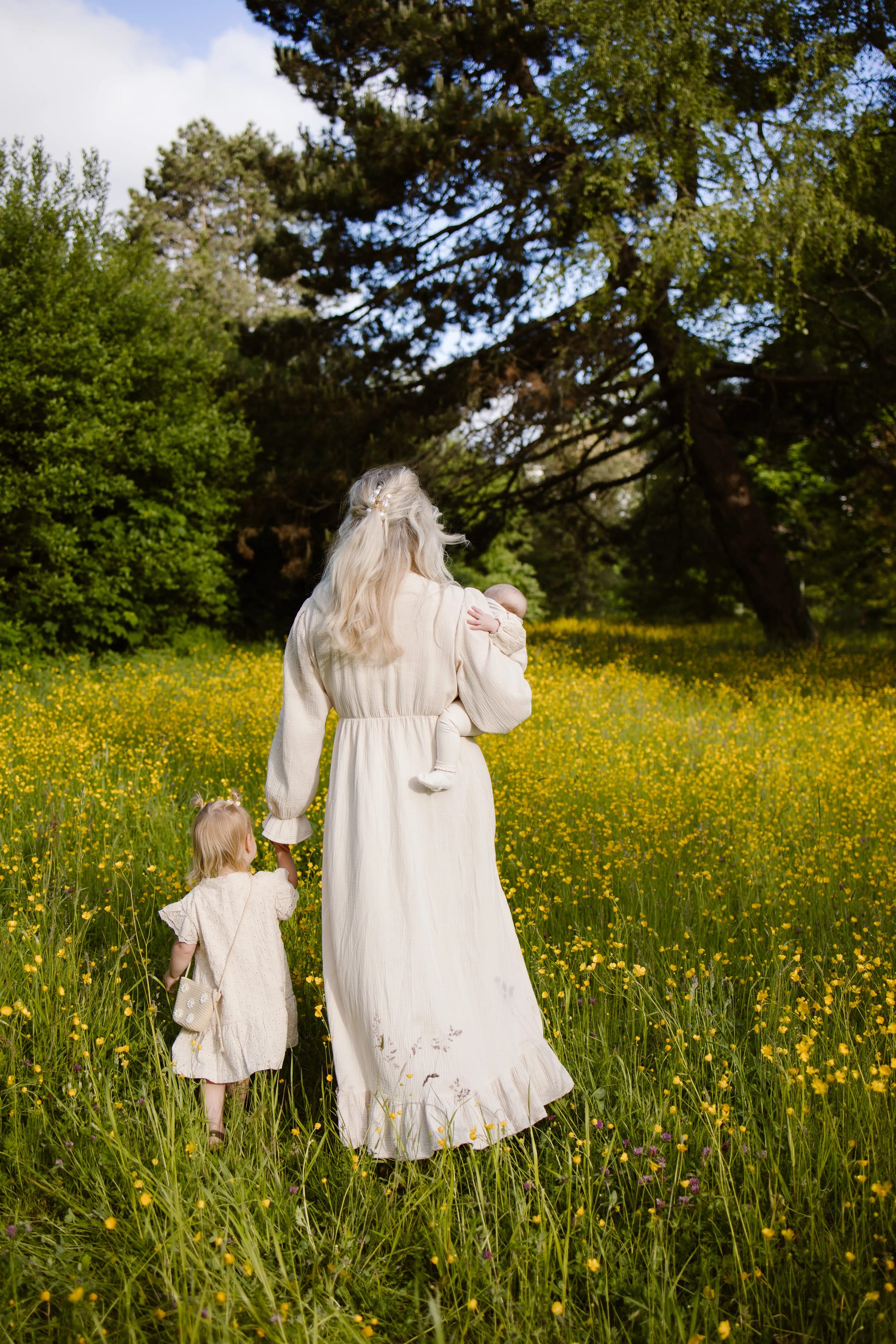 Een vrouw met lang, grijs haar in een wit, vloeiend kleed loopt hand in hand met een klein meisje door een bloeivelden gevuld met gele bloemen, terwijl ze een baby op haar heup draagt.