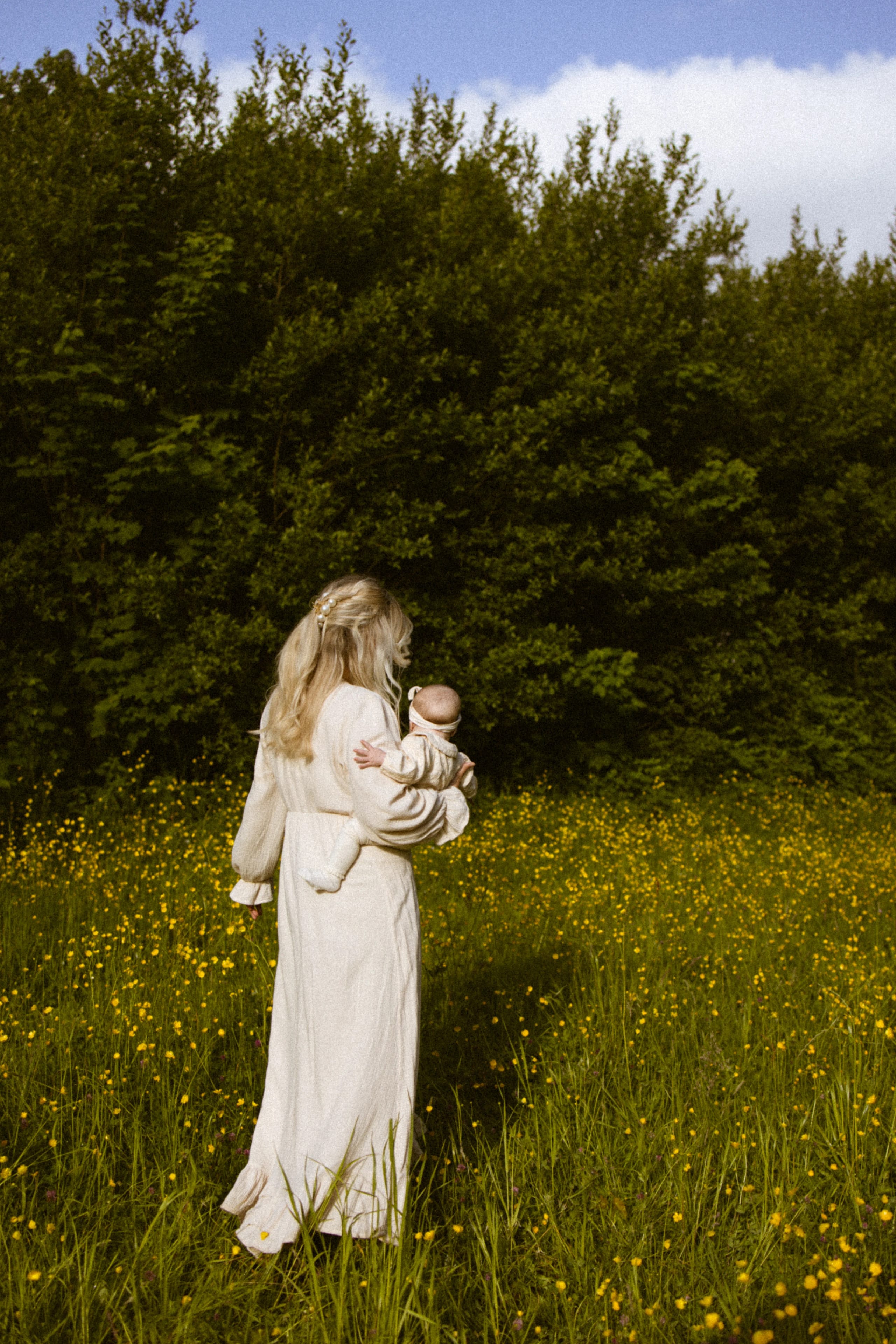 Een vrouw in een witte omslagdoek houdt een baby vast terwijl ze door een bloeiende veld met gele bloemen loopt, omringd door groene bomen en een heldere lucht.