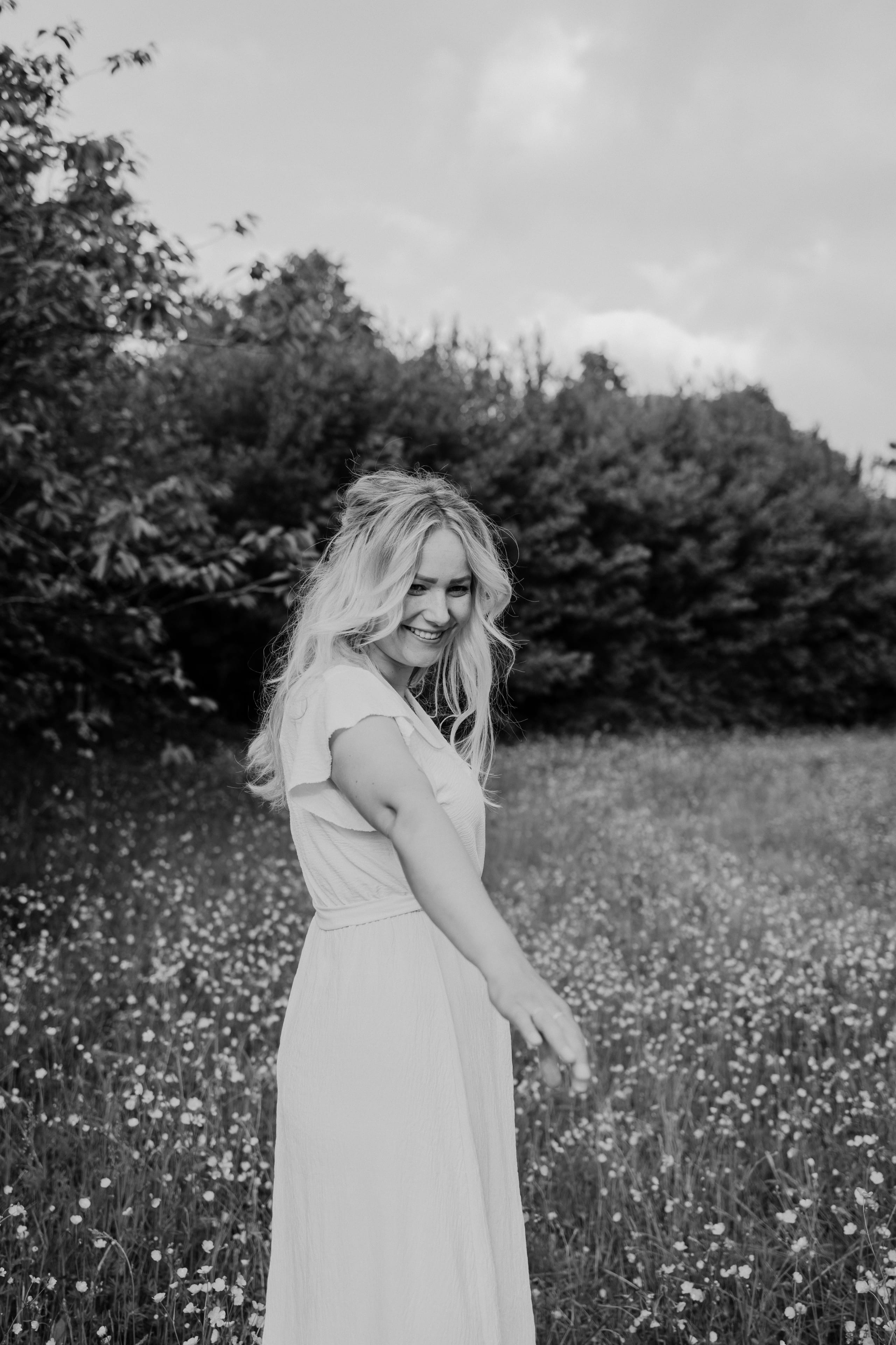 Jonge vrouw met lange, golvende haren in een witte jurk, die glimlacht terwijl ze zich omdraait in een bloeiend veld met bloemen en groene struiken op de achtergrond.