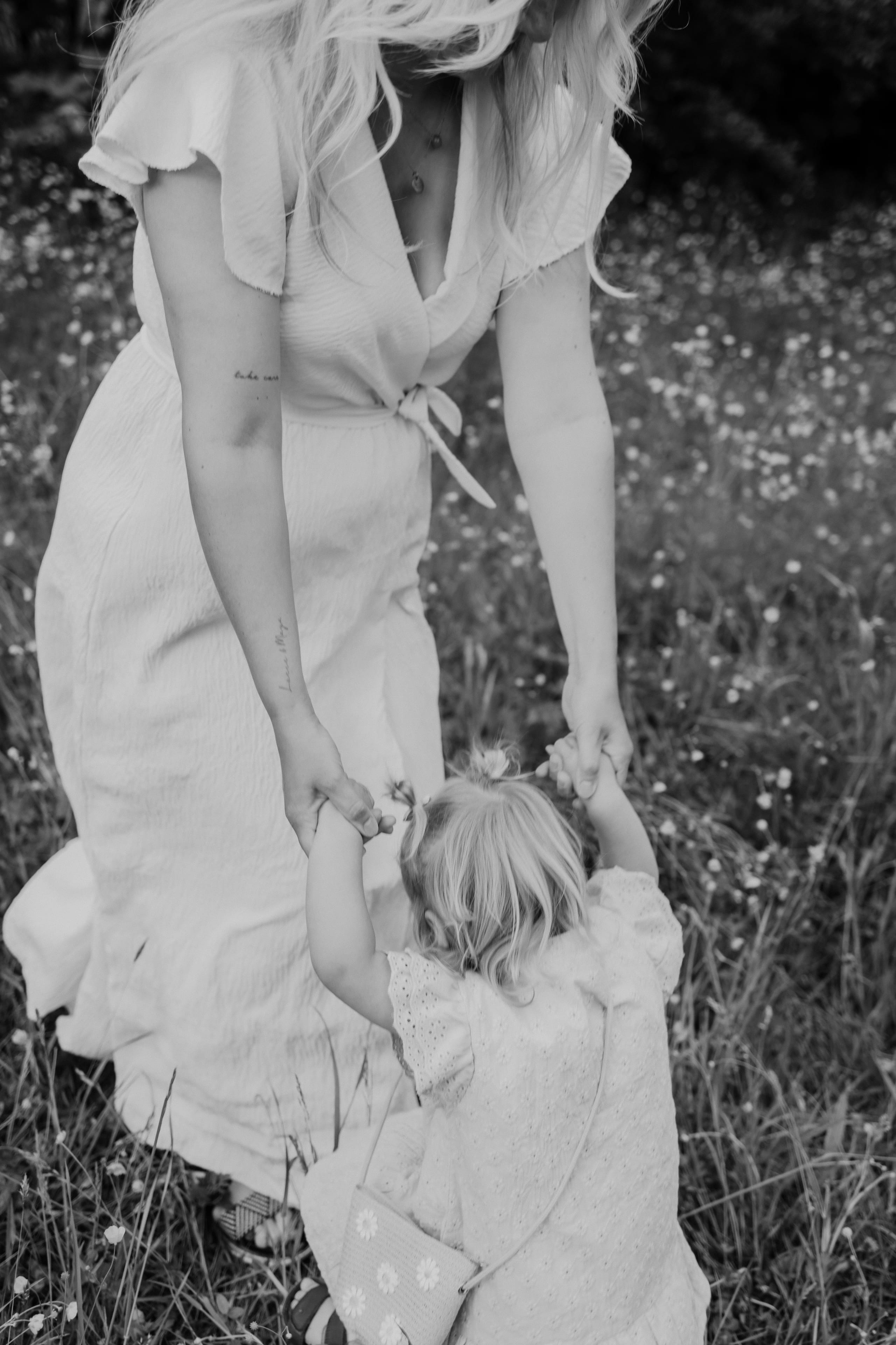 Een moeder helpt haar kleine dochter rechtop staan in een bloeiende groene veld, terwijl ze elkaar handjes vasthoudt. De moeder draagt een stijlvolle, lichte jurk, terwijl het meisje een schattige, lichte outfit met stippen aan heeft.